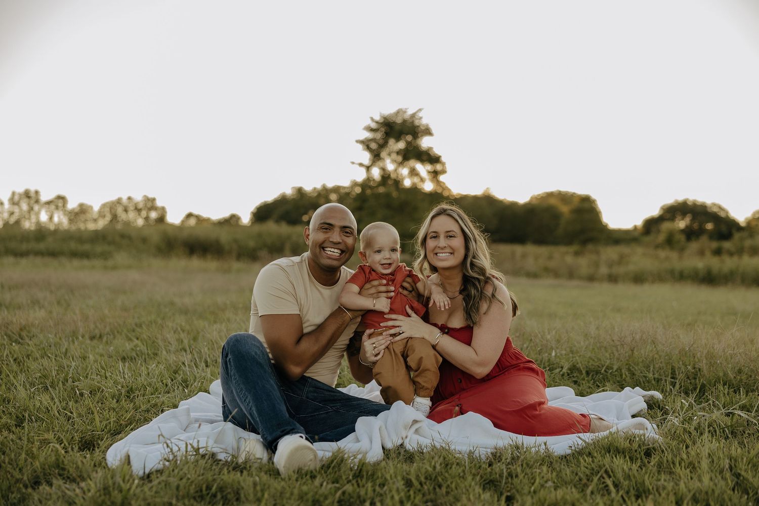 Family relaxes together on white blanket during outdoor sunset photoshoot.