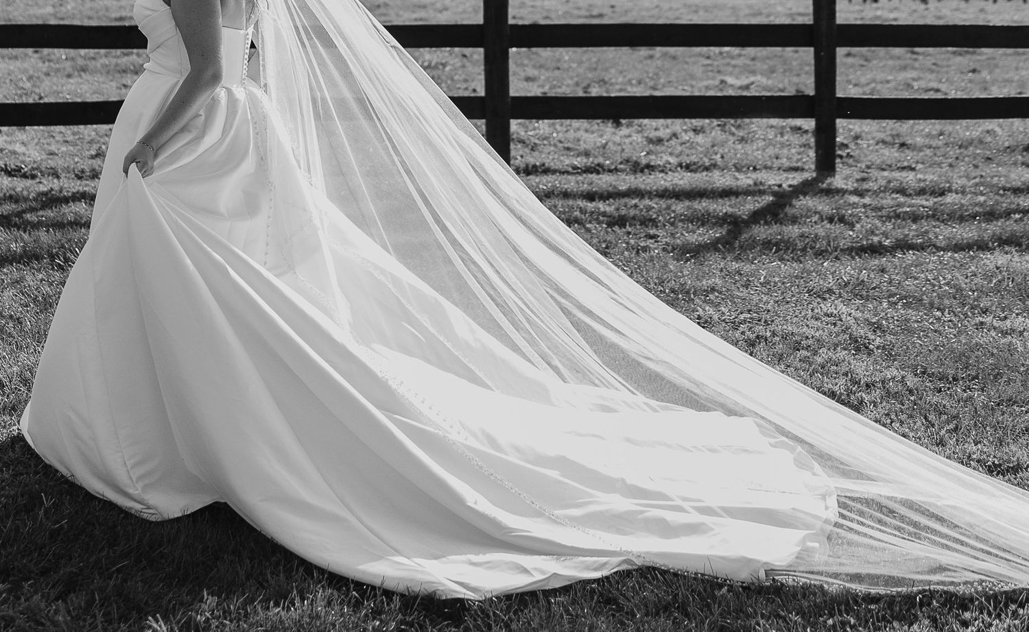 Flowing white wedding dress train trailing across grass with wooden fence in background.