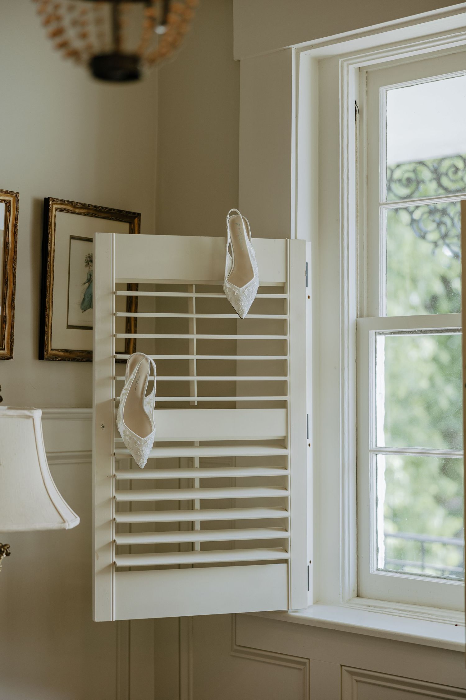 White bridal heels hanging on plantation shutters near a sunlit window in an elegant room.