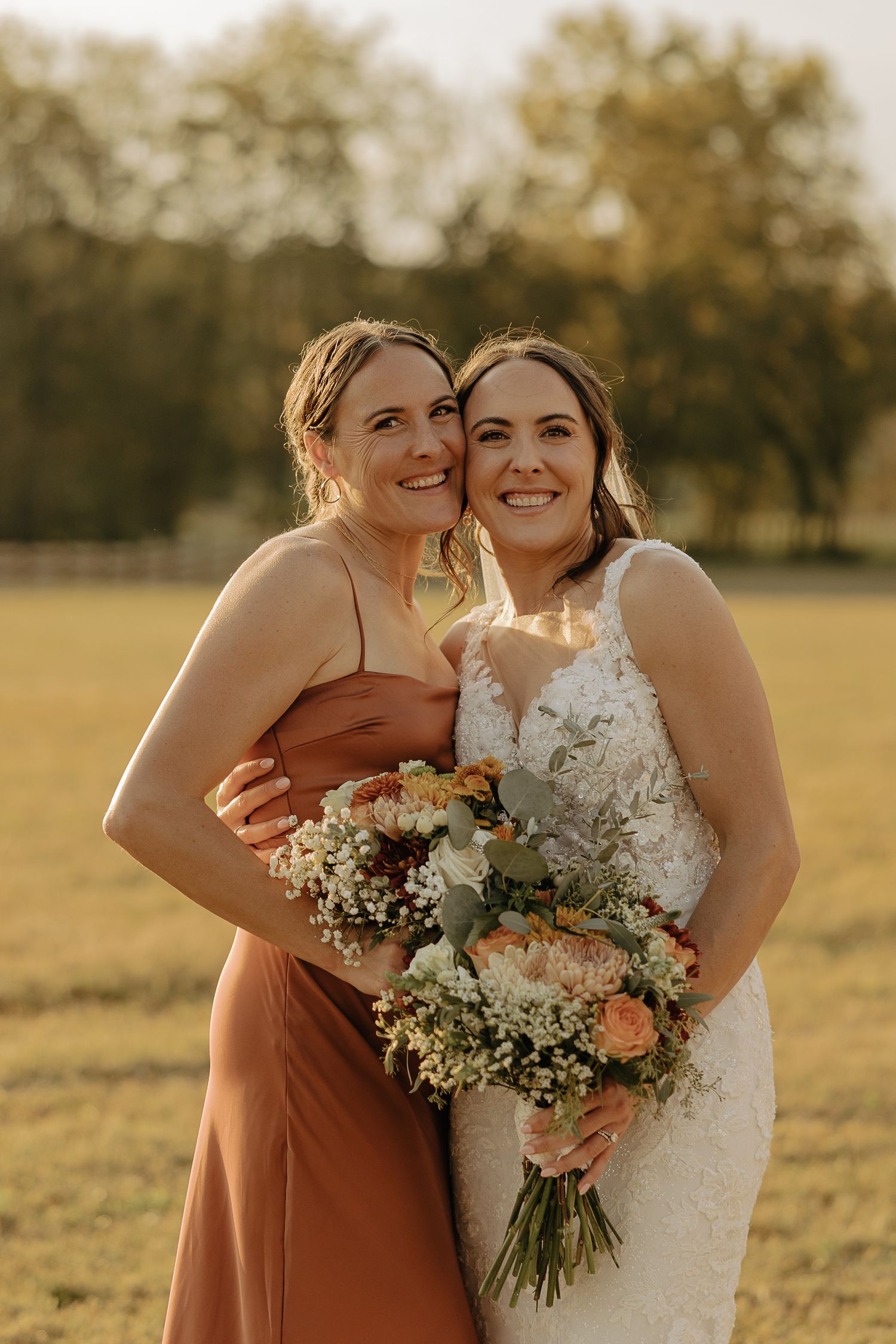Wedding party members in rust-colored dresses embrace during outdoor golden hour photography.