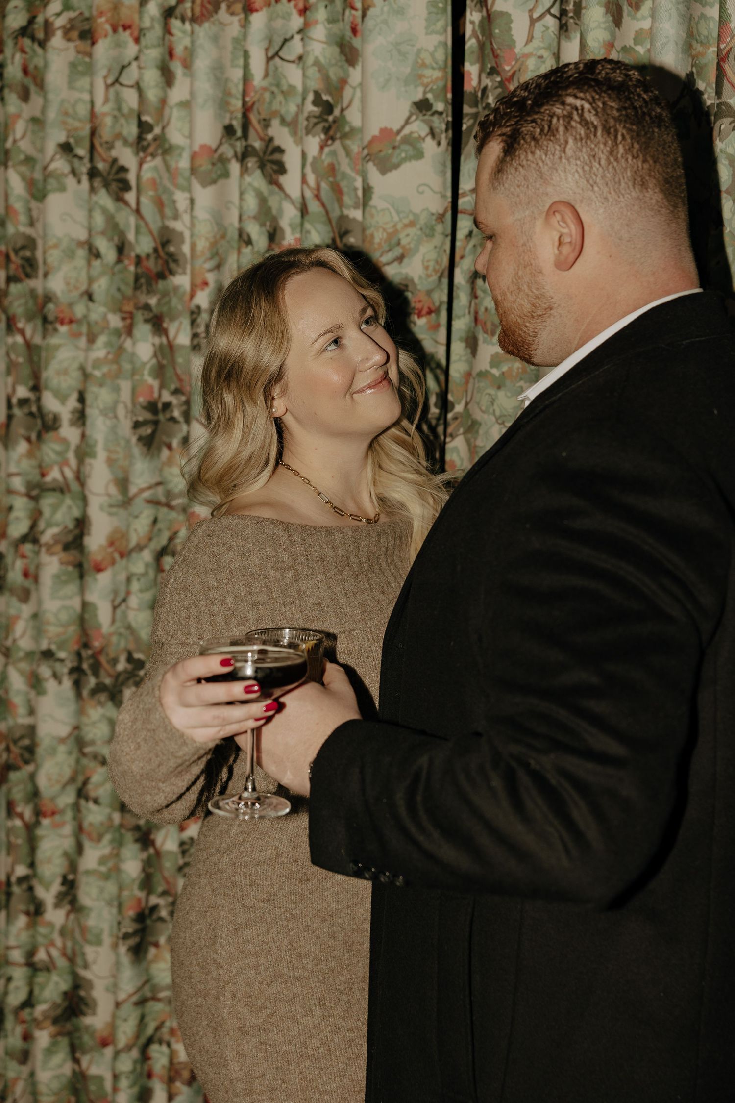 A couple shares drinks in an intimate setting with floral curtains in the background.