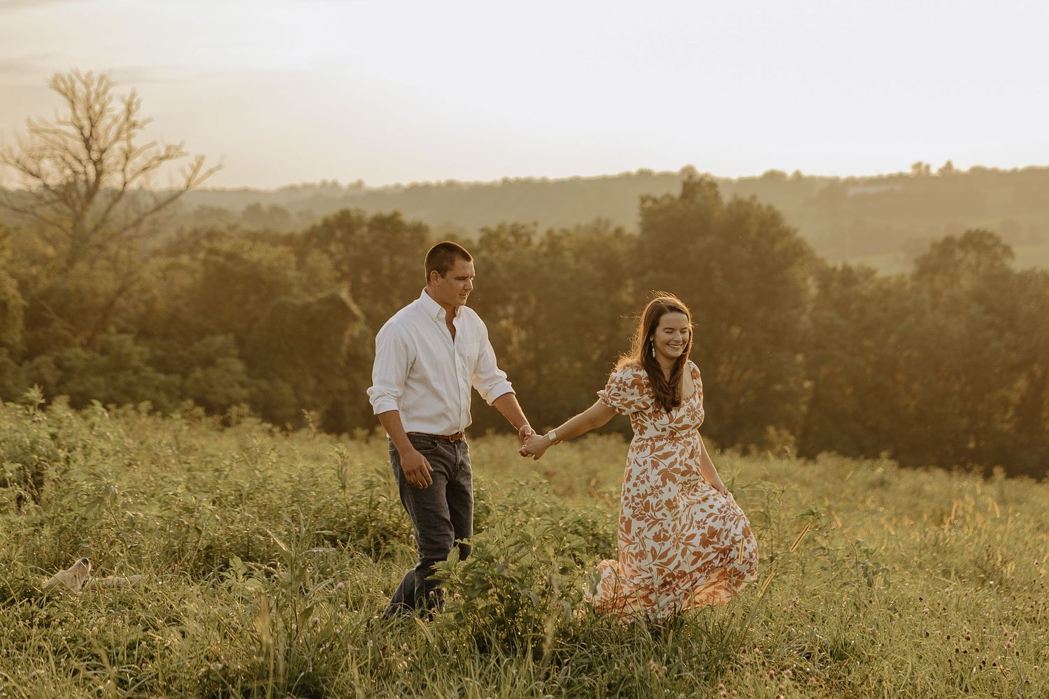 A couple walks through a golden meadow at sunset holding hands in a romantic outdoor setting.