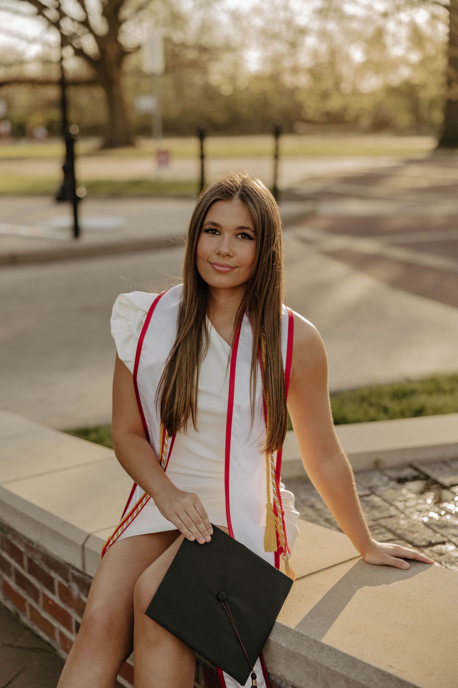 Graduate holding mortarboard cap while sitting on brick ledge during sunset photo session.