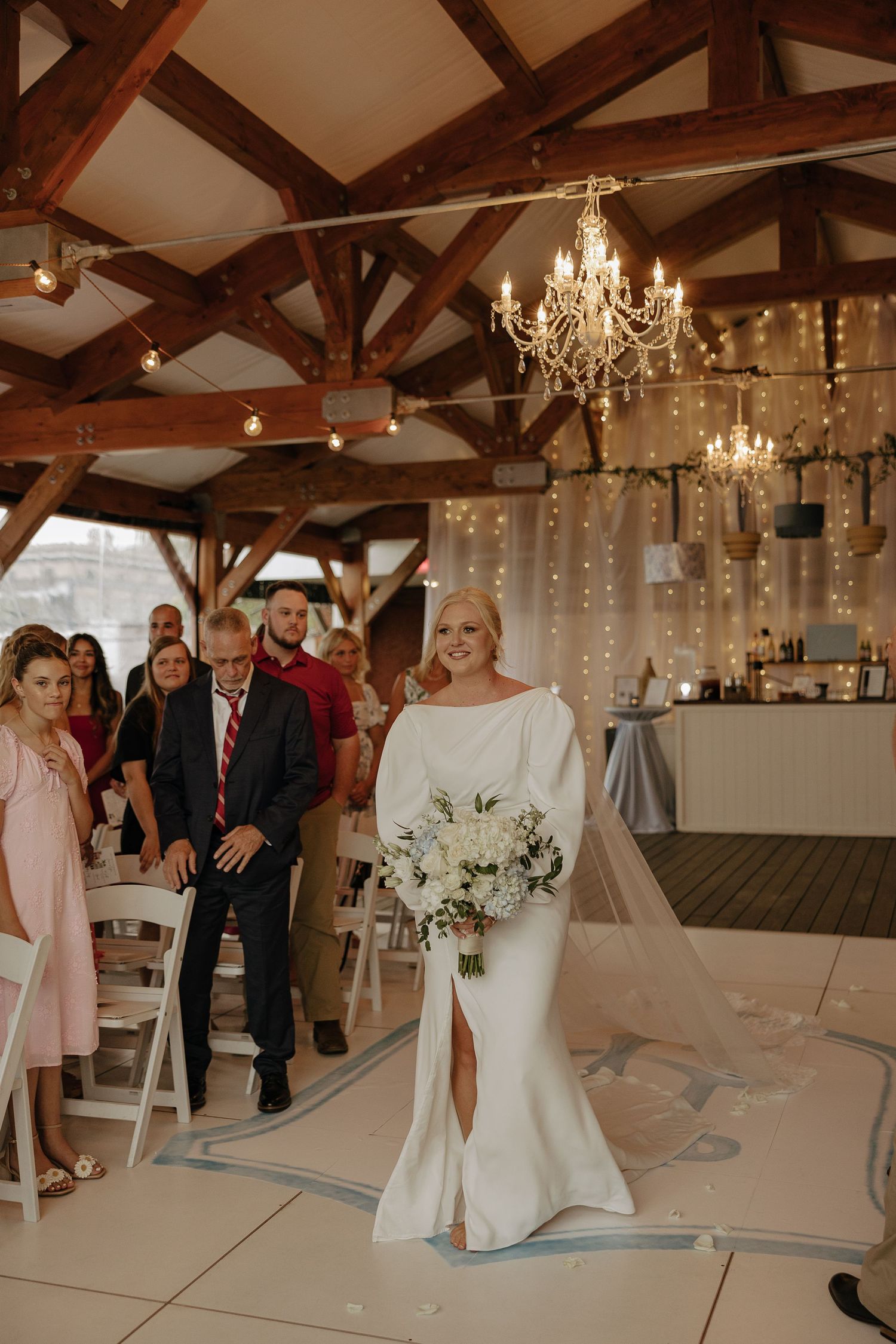 Wedding reception venue with wooden beams, chandeliers, and elegant white decor.
