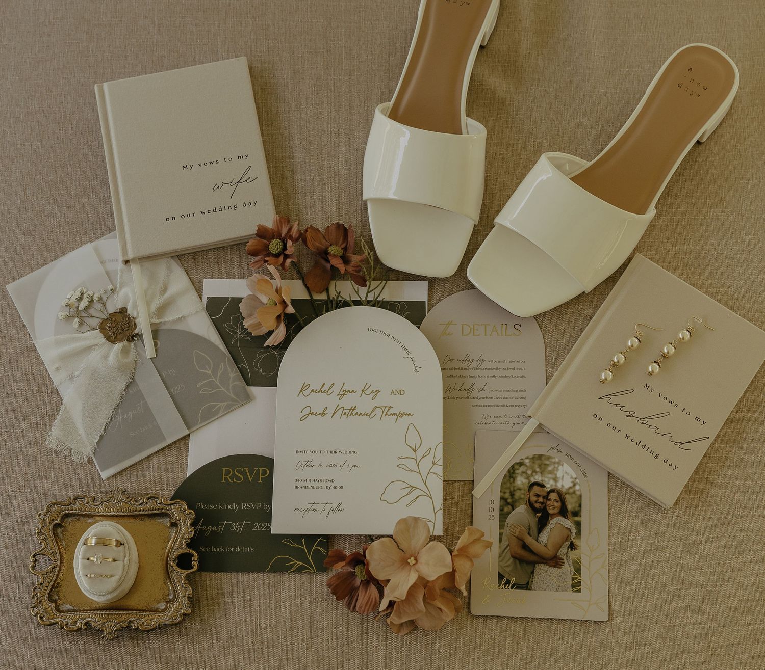 Elegant wedding stationery collection featuring cream-colored invitations, envelopes, and dried floral accents laid out on beige surface.