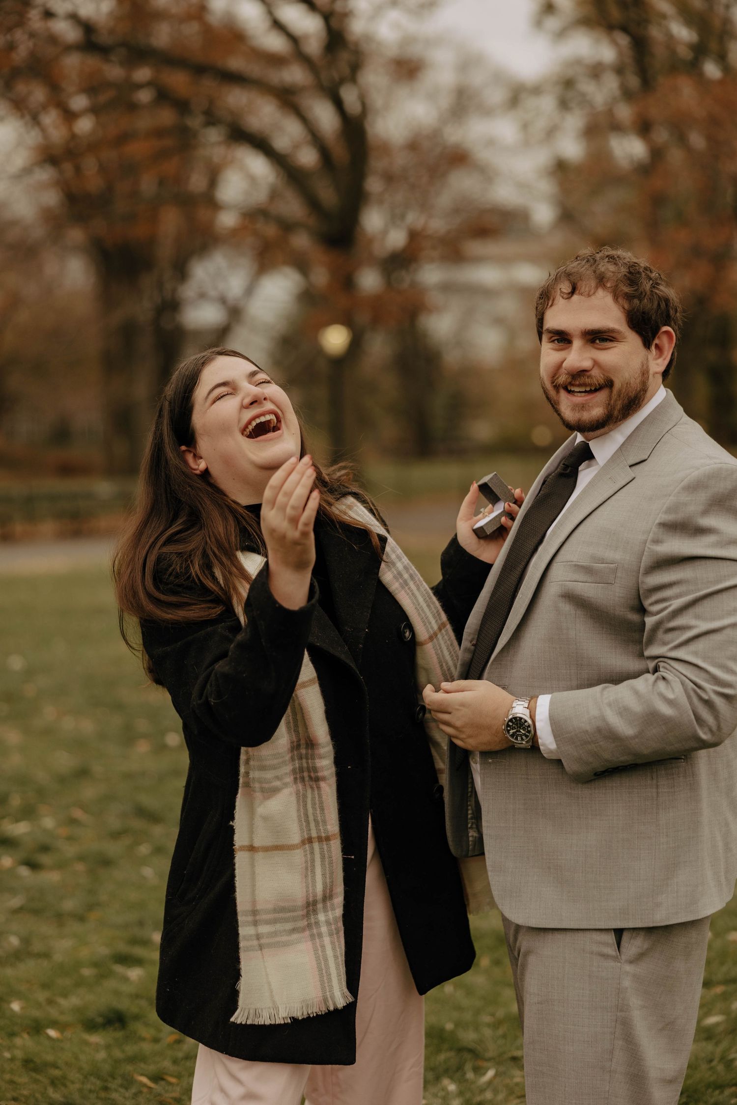 A joyful autumn engagement photo showing a couple laughing together in a park with fall foliage in the background.