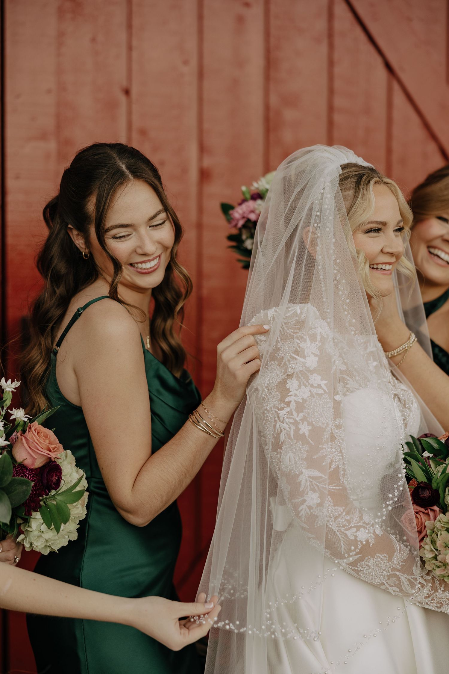 Bride in white lace veil and bridesmaids in emerald green dresses share joyful laughter during wedding photos.