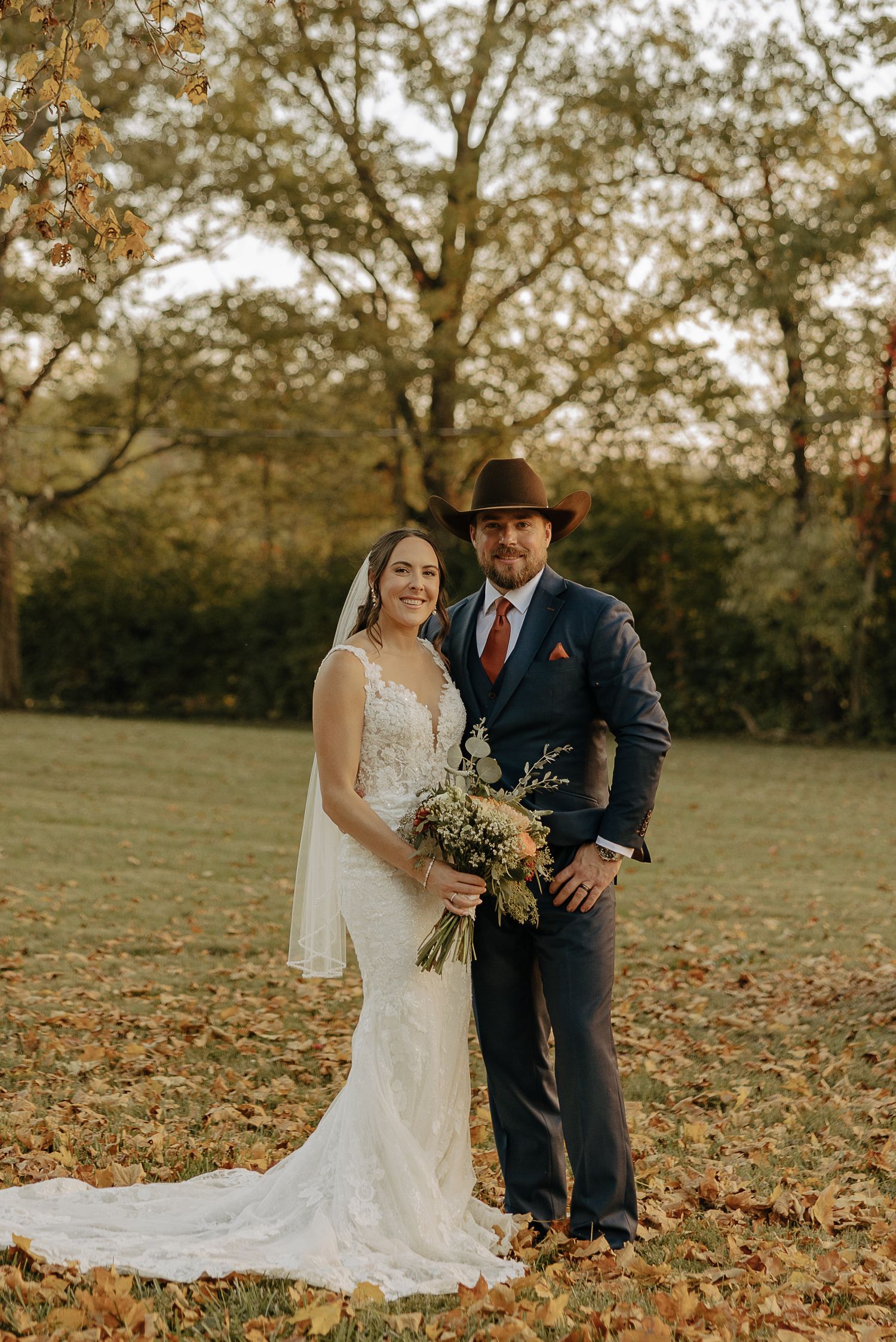 A couple poses among fallen autumn leaves in a rustic outdoor wedding setting.