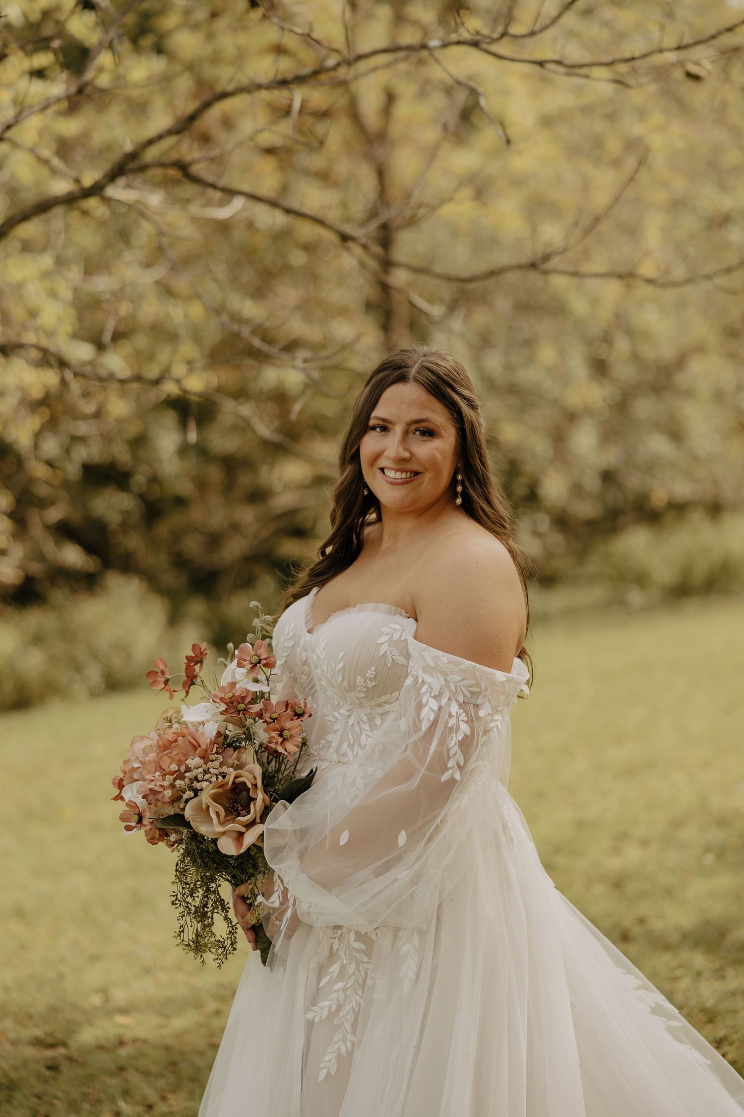 Bride in an off-shoulder lace wedding gown poses outdoors with pink floral bouquet.