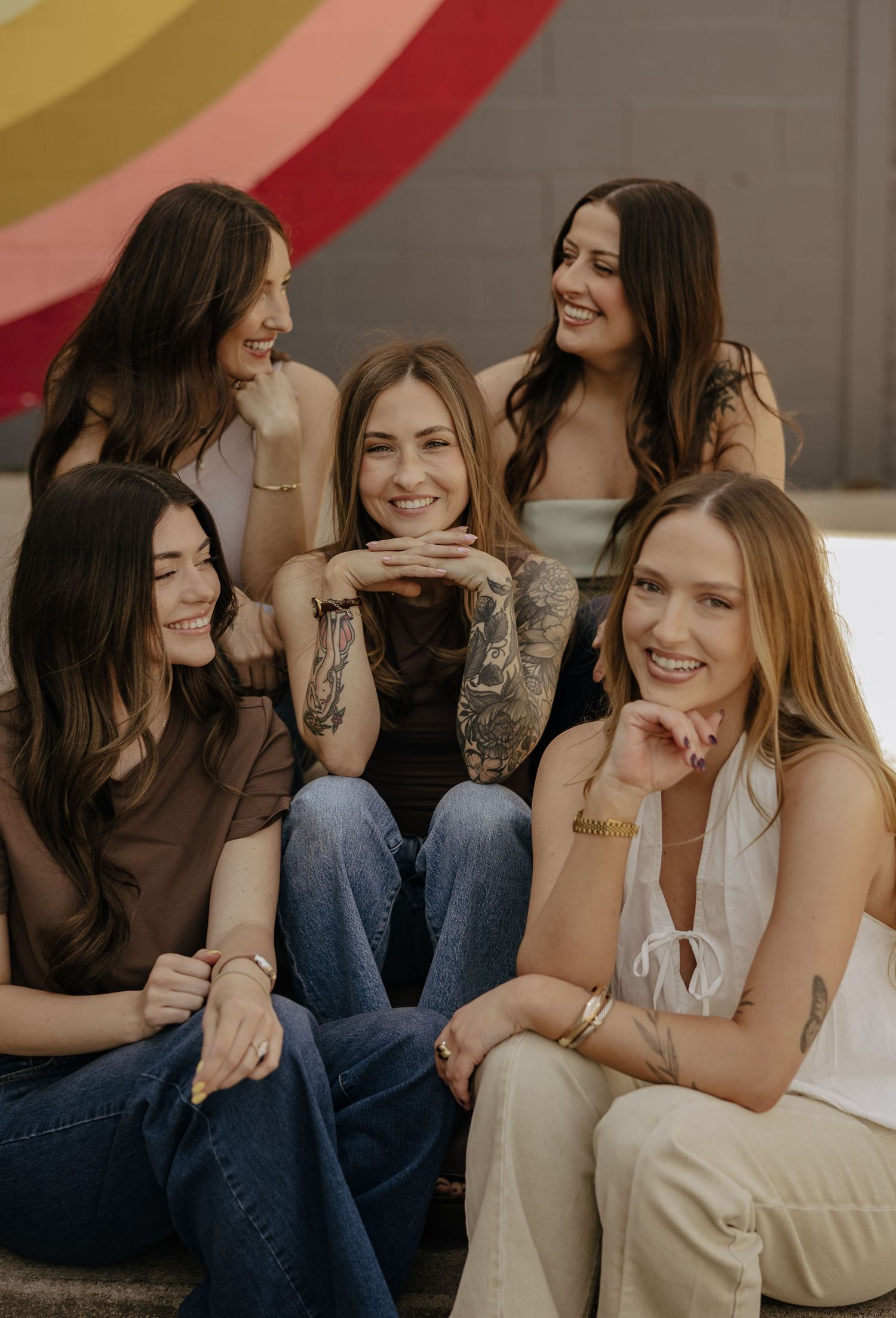 Five smiling women with tattoos pose together in front of a colorful rainbow mural outdoors.