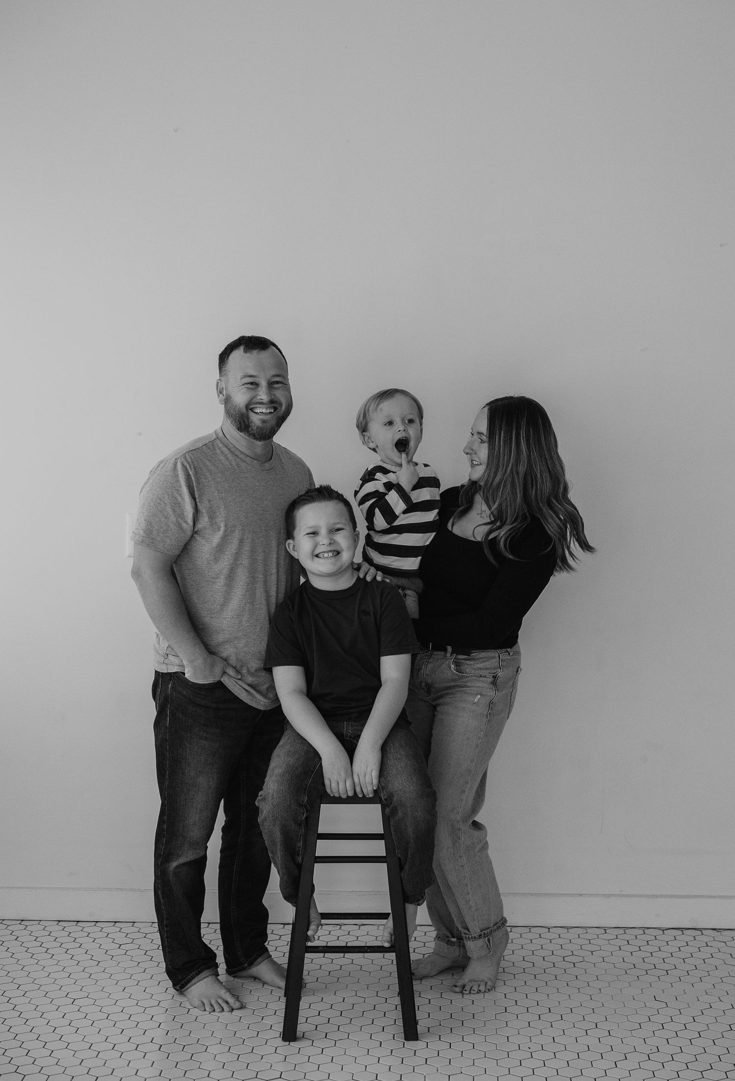 A casual black and white family portrait against a plain wall with a wooden stool as prop.