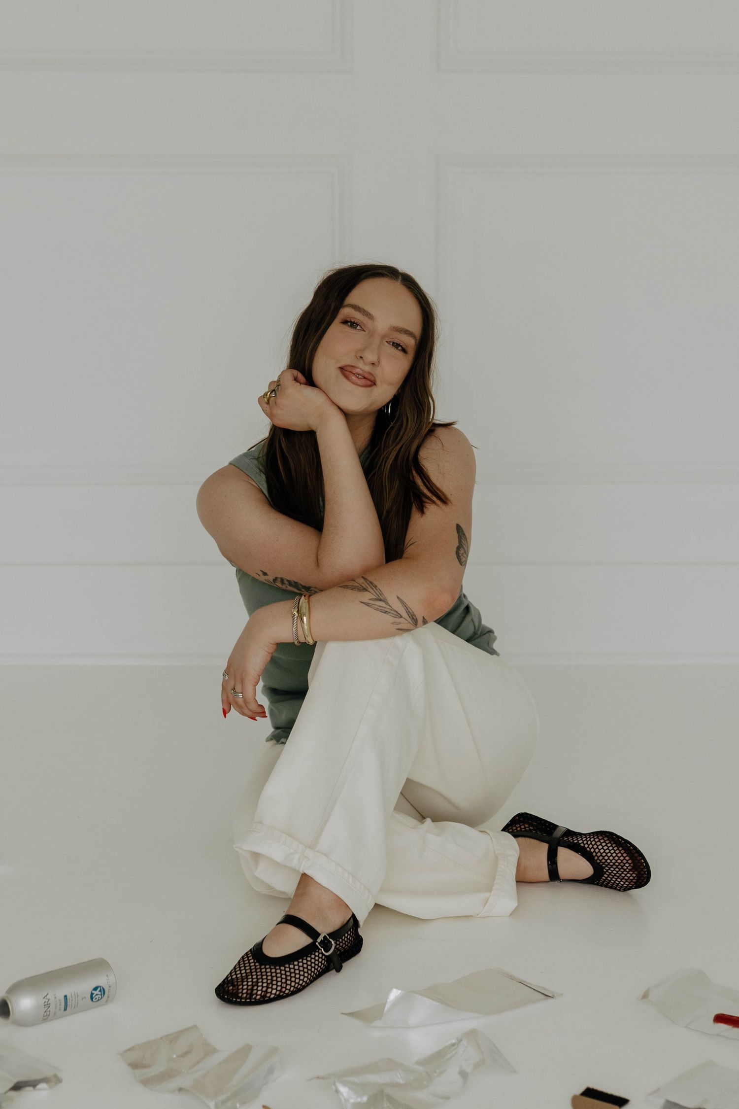 A person in white clothing sits casually on the floor with scattered papers and beauty products.