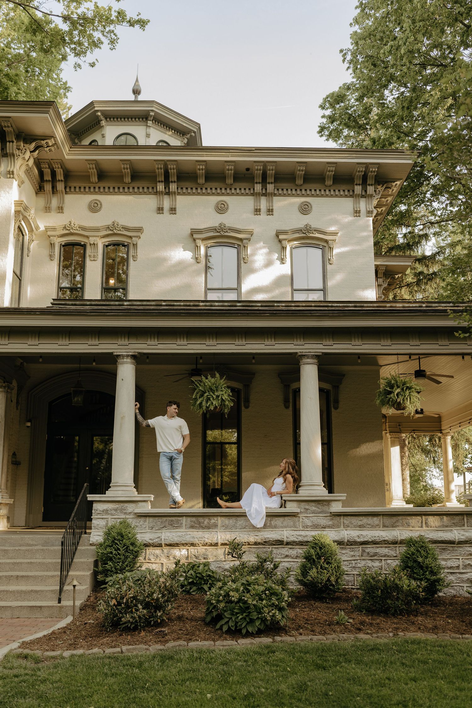 Couple posing on the porch of a grand historic two-story home with white columns and ornate architectural details.