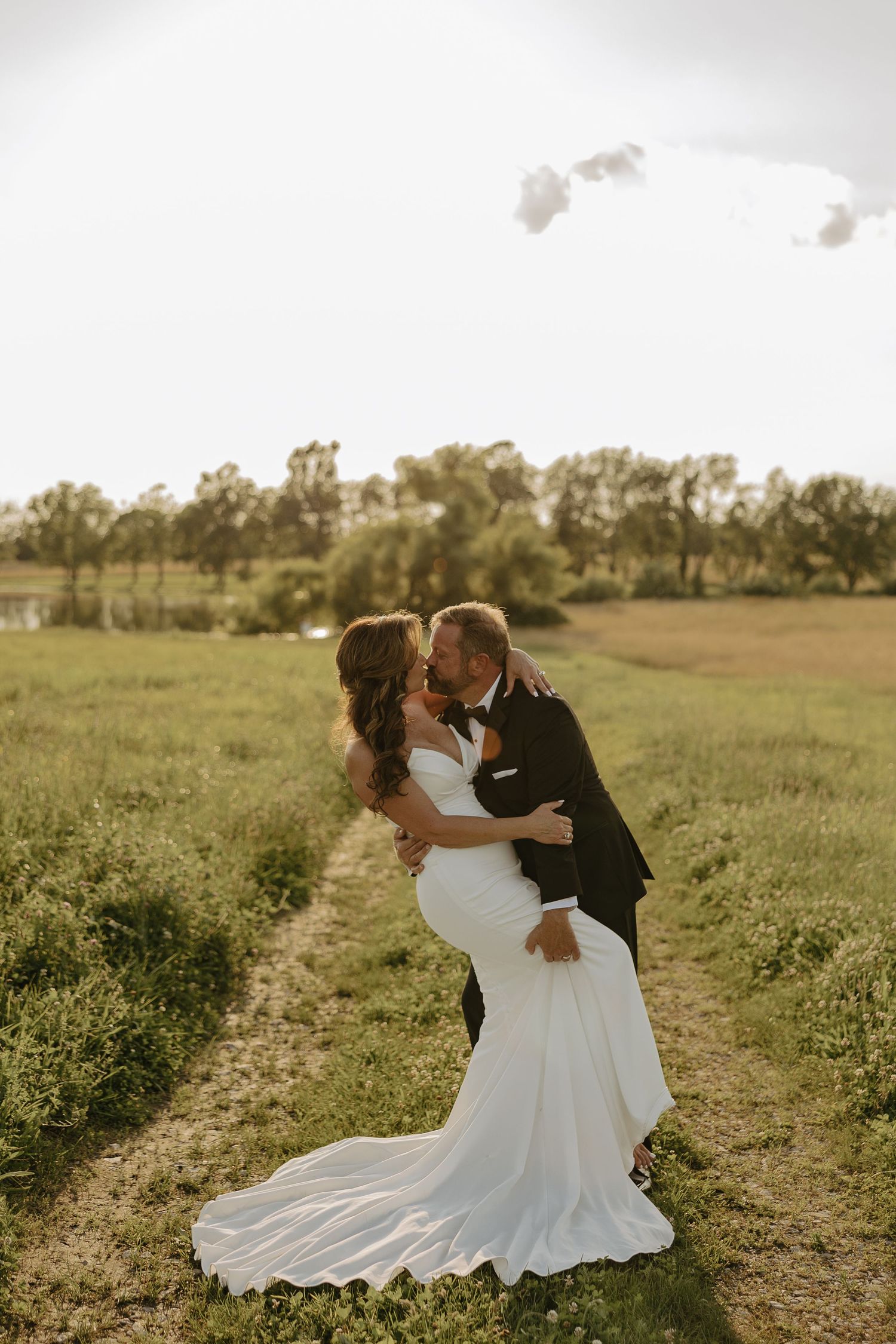 A couple shares a romantic kiss in a golden field at sunset while the bride's white dress train flows behind her.