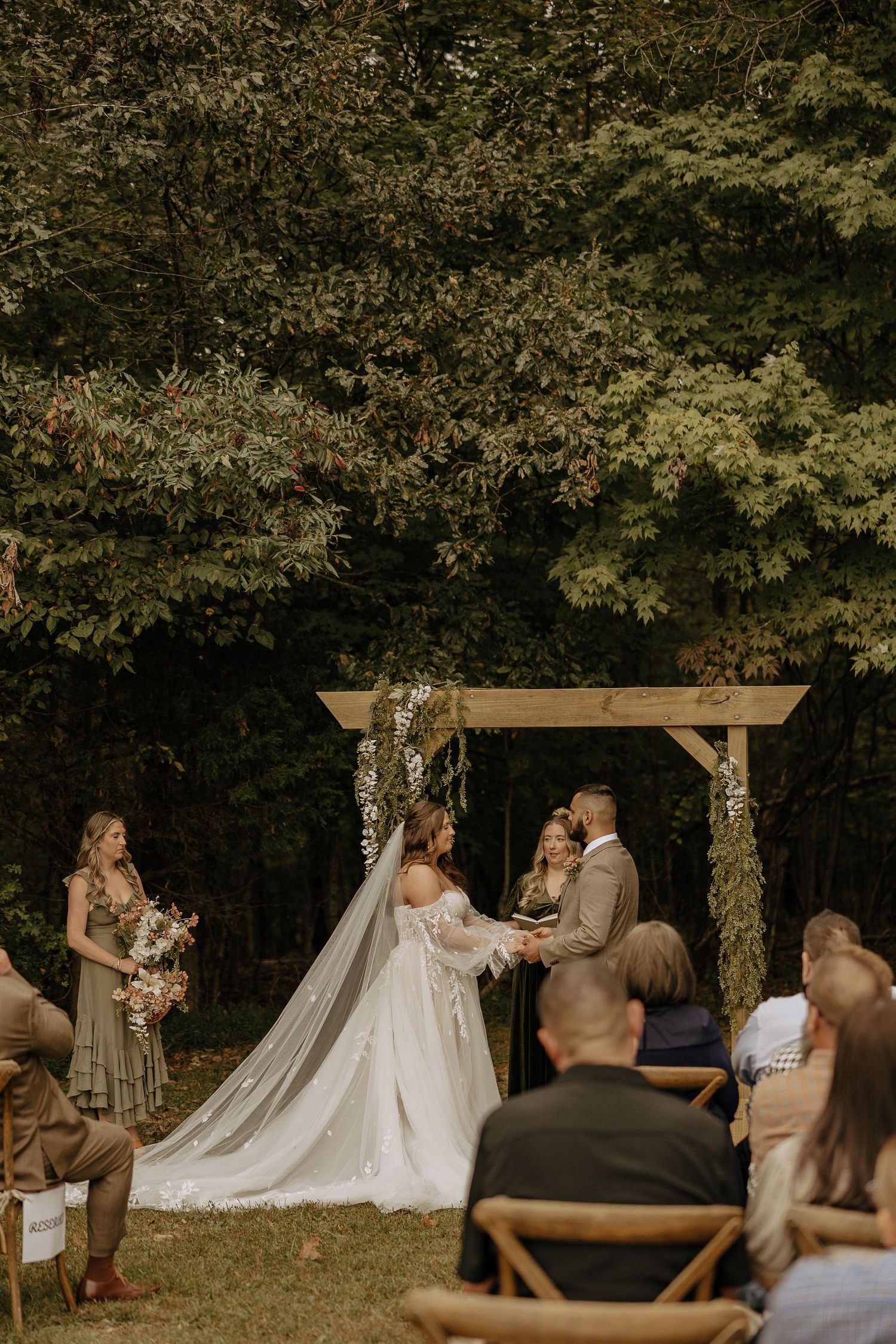 Outdoor wedding ceremony under wooden pergola decorated with floral arrangements and greenery.