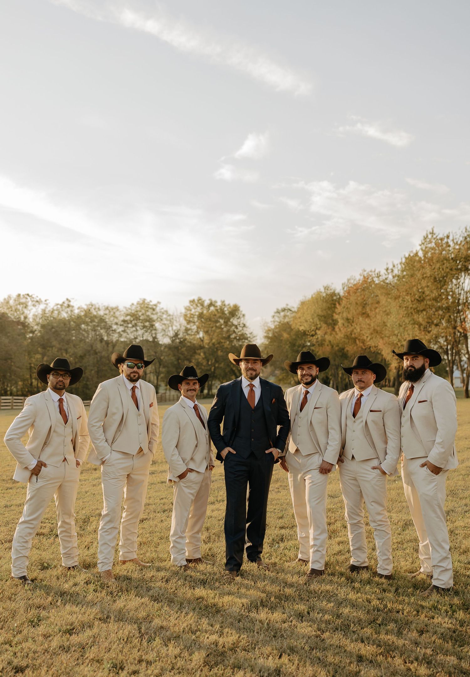 Groomsmen in white suits and black cowboy hats line up in a field at sunset.