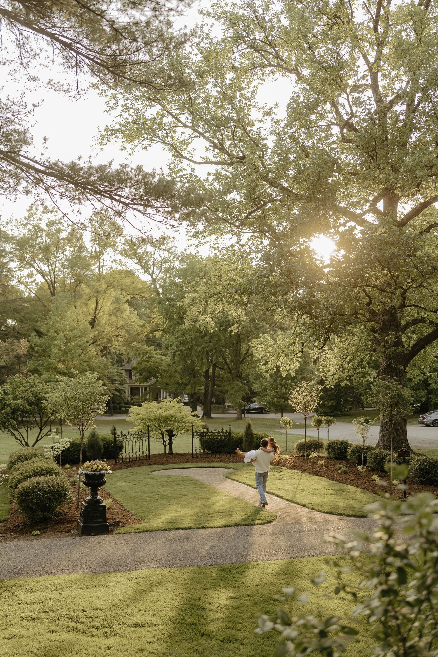 Couple embracing on a curved driveway surrounded by lush green trees and manicured lawn at golden hour.