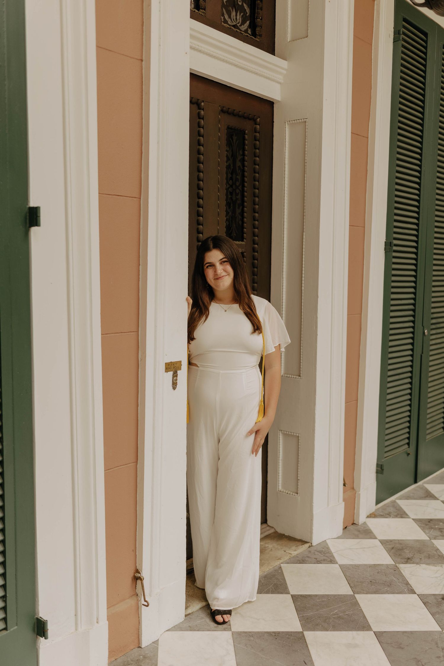 A graduate poses between white columns and green shutters of a historic building.