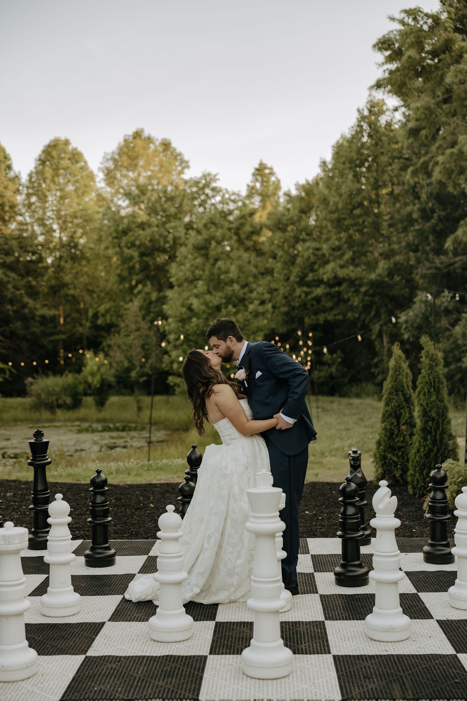 A couple shares a romantic moment on a giant outdoor chess board surrounded by life-sized white chess pieces at sunset.
