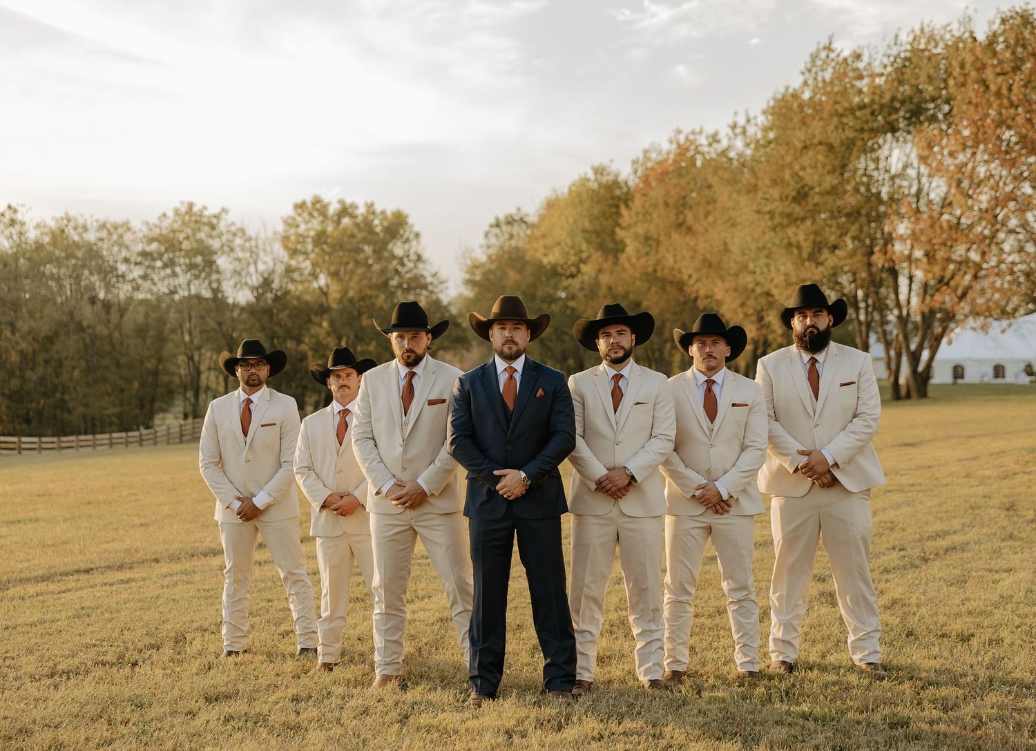 Wedding party members in white suits and black suits pose together outdoors.