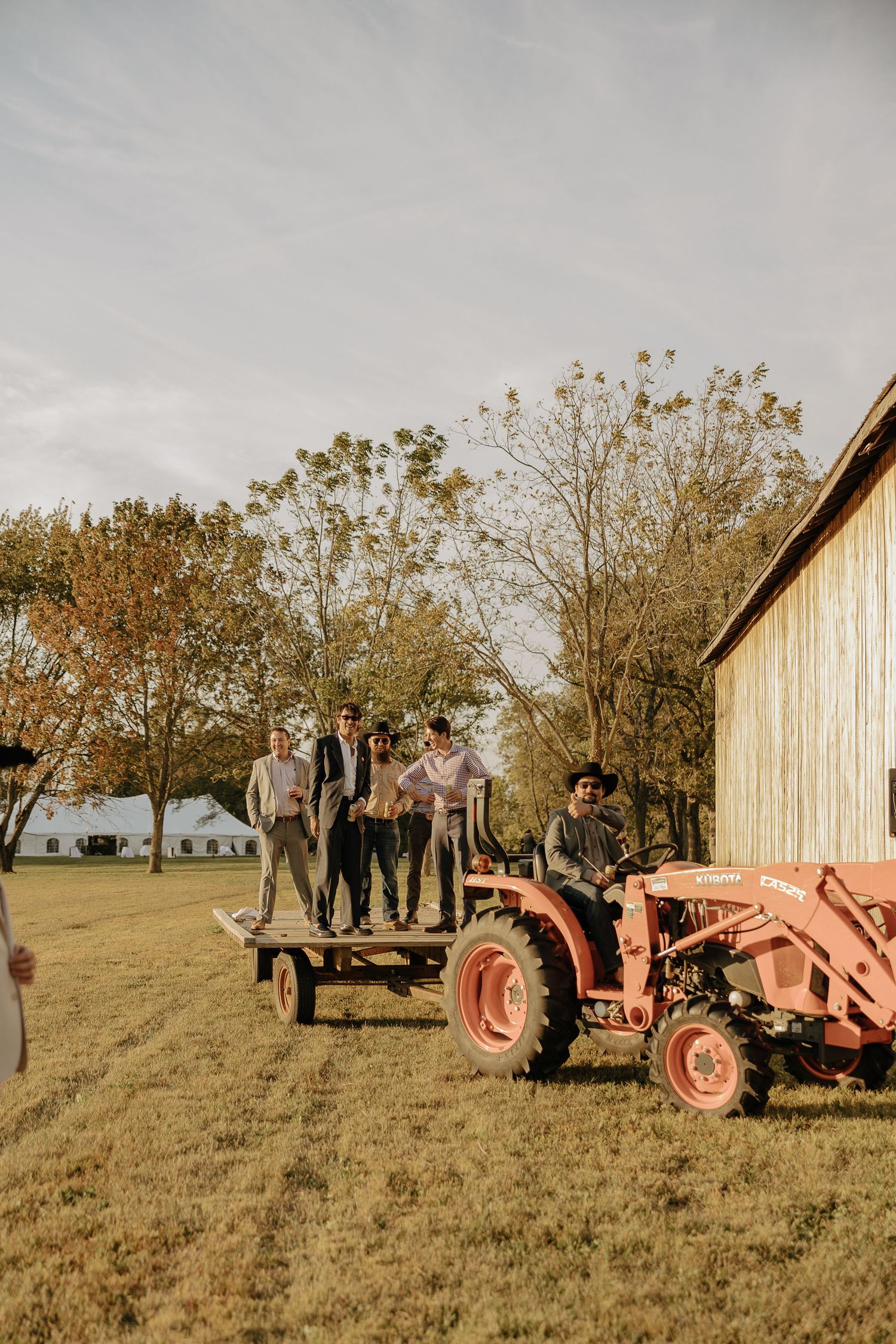 Wedding party members pose on a wagon being pulled by a vintage red tractor near a rustic barn.