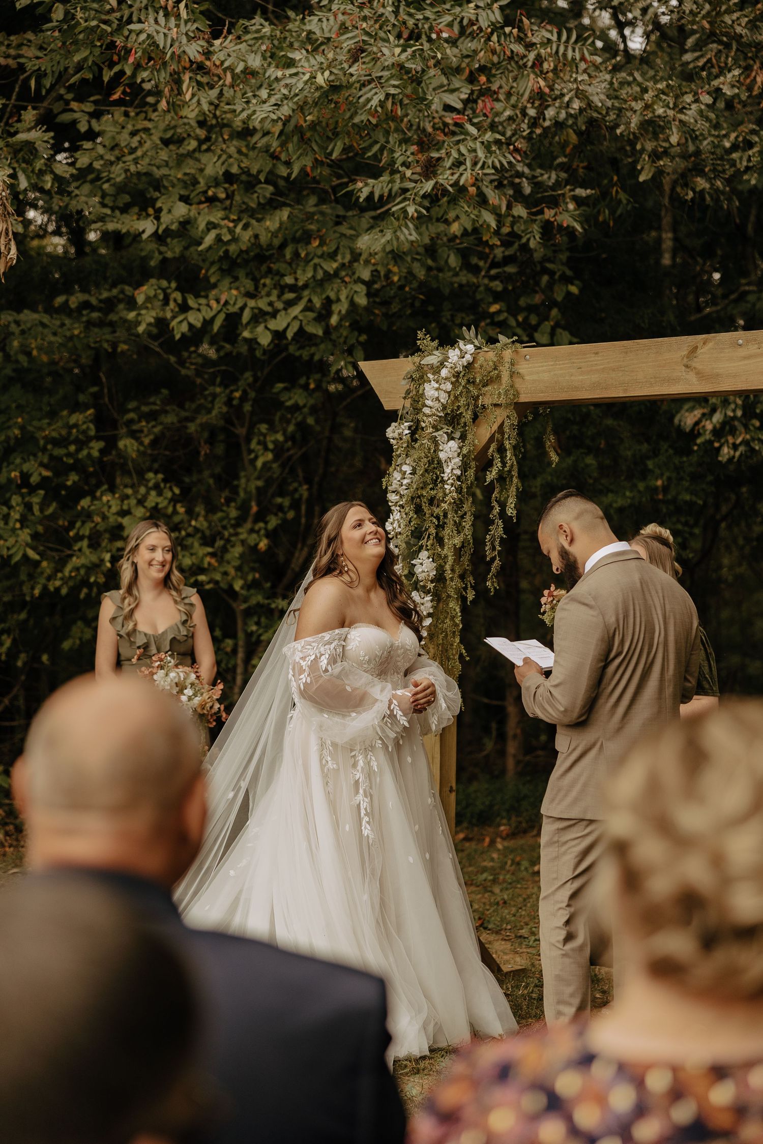 An outdoor wedding ceremony takes place under trees with wooden chairs and floral decorations.
