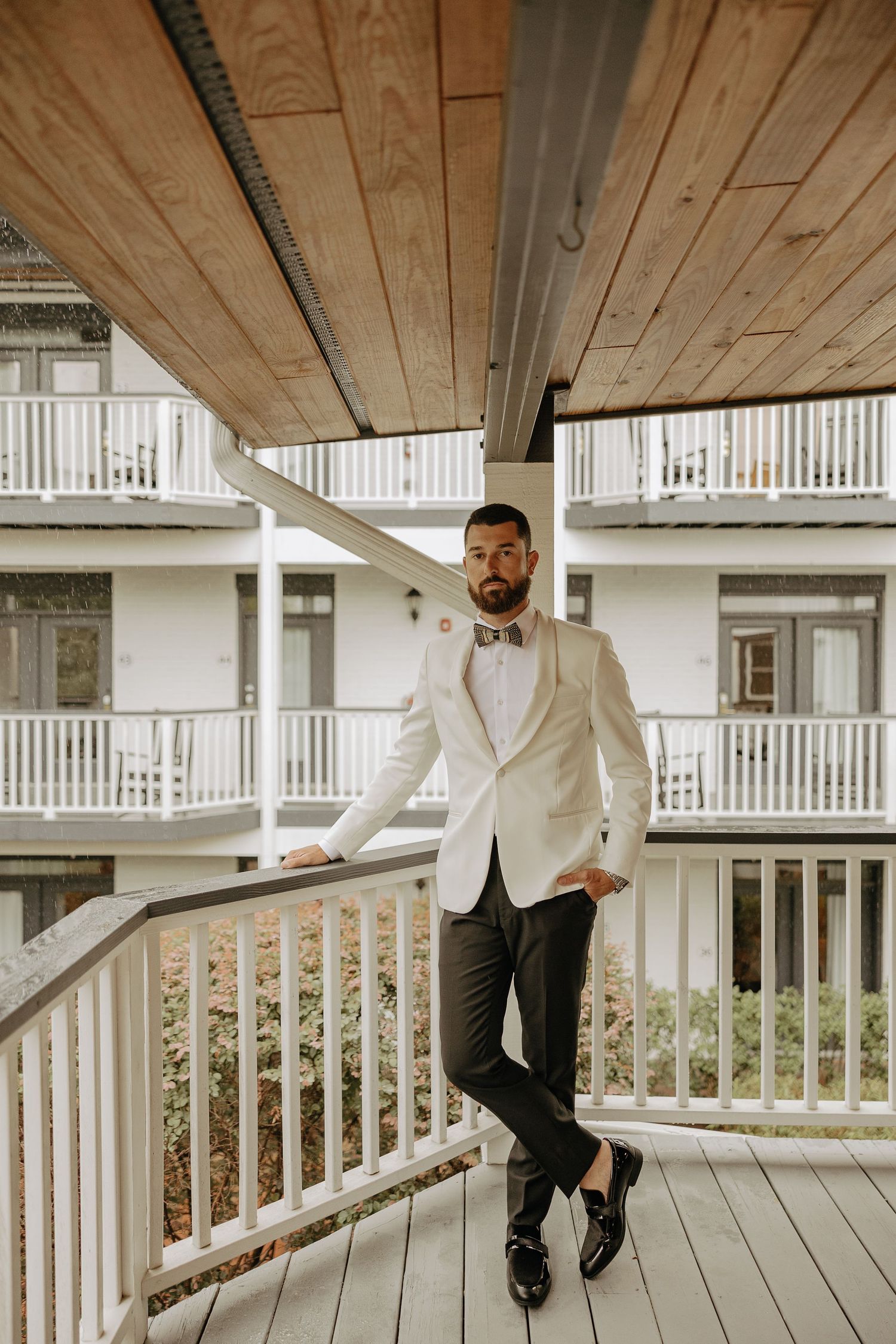 A person in a white blazer and dark pants poses casually on a wooden deck with white railings.