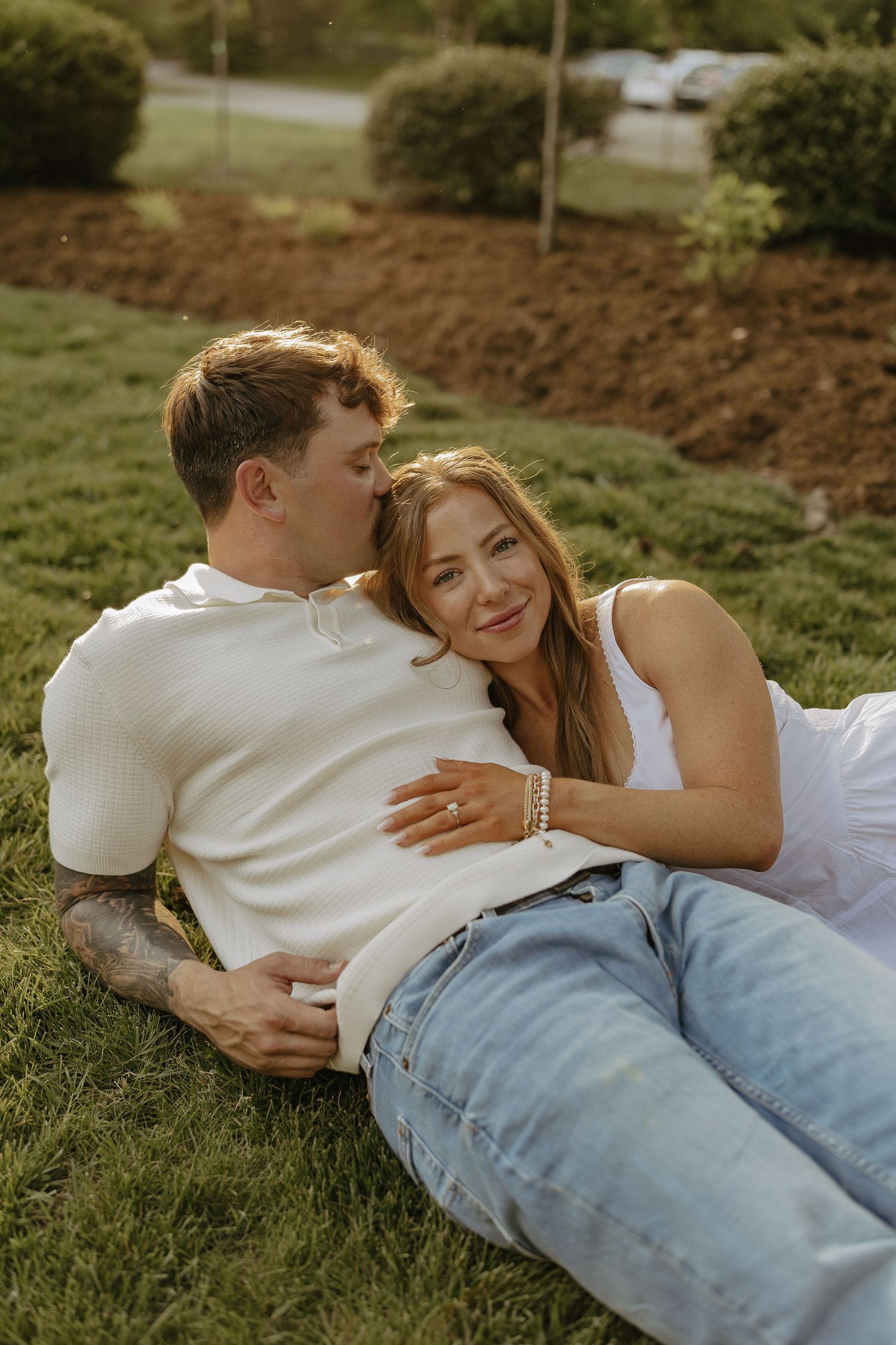 Couple relaxing on grass, man kissing woman's head, both in white tops and jeans outdoors.