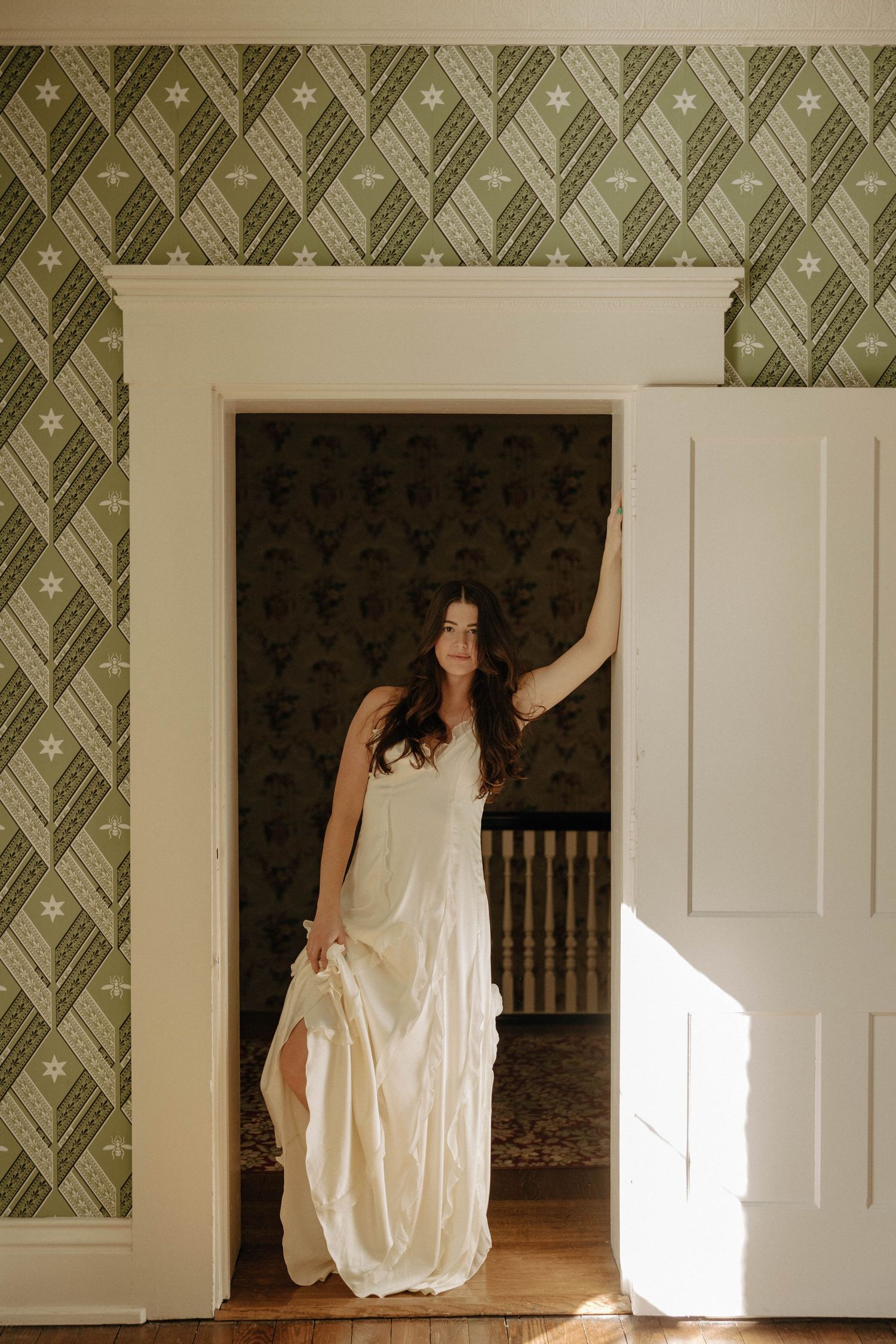 A white wedding dress emerges from a doorway against green patterned wallpaper in vintage interior setting.