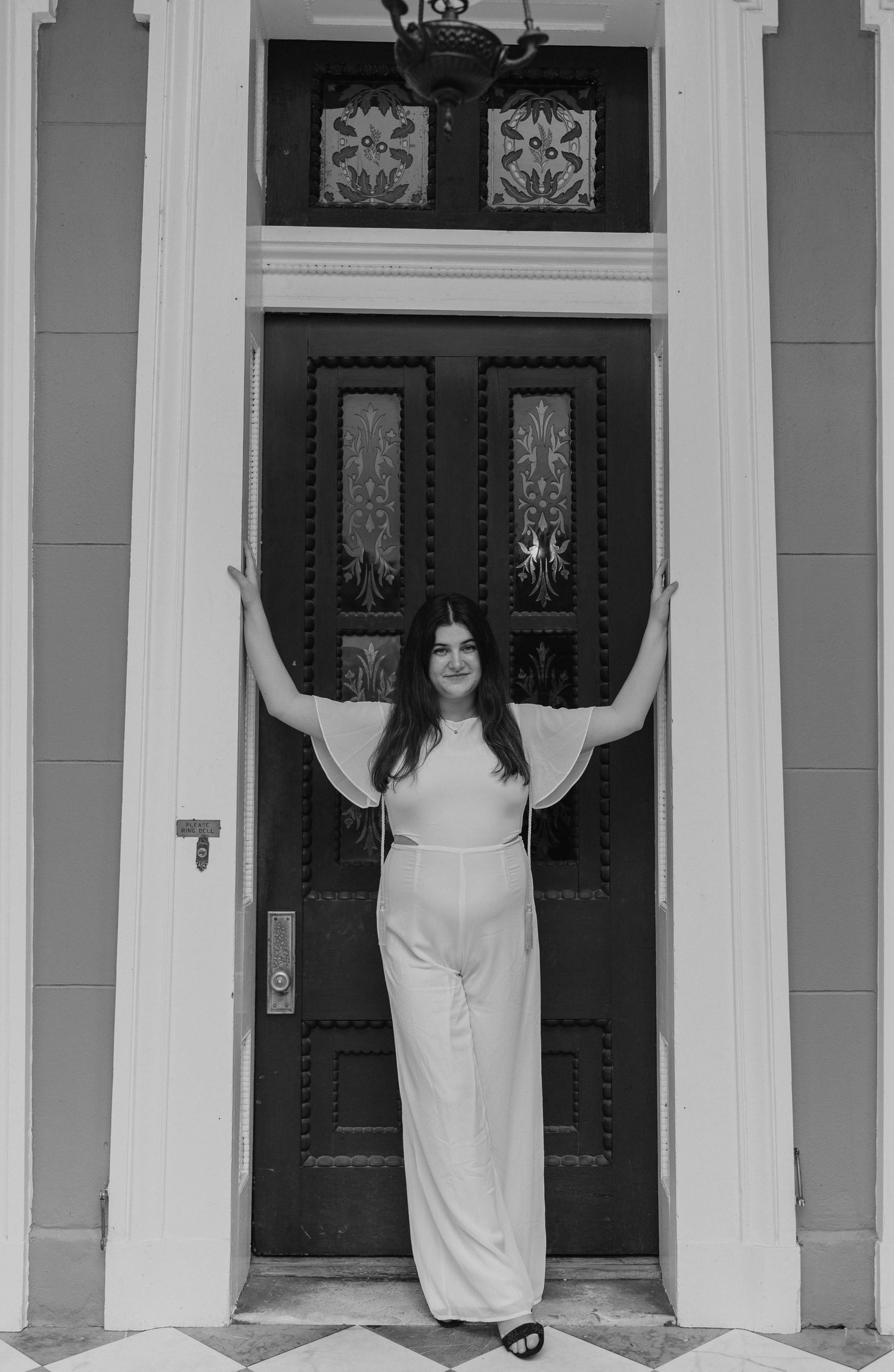 Graduate in white dress poses playfully between white columns of a classic building entrance.