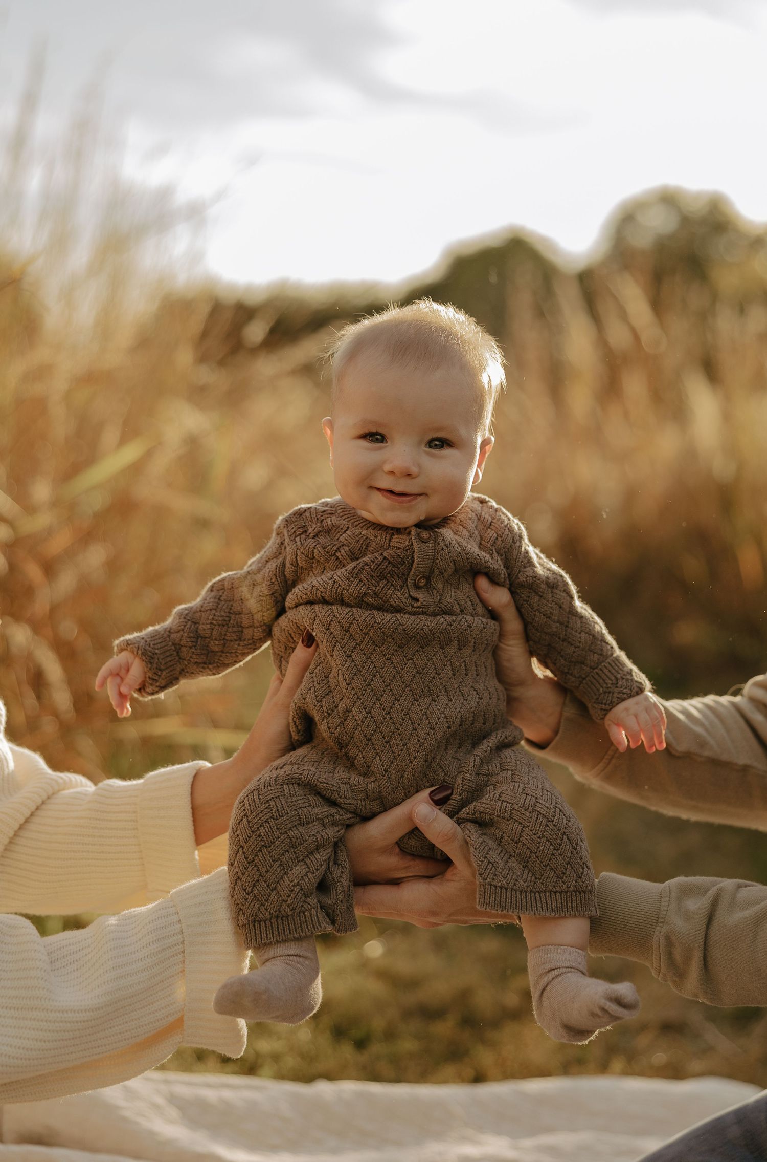 A baby in a brown knit outfit smiles while being held up against a warm autumn background.