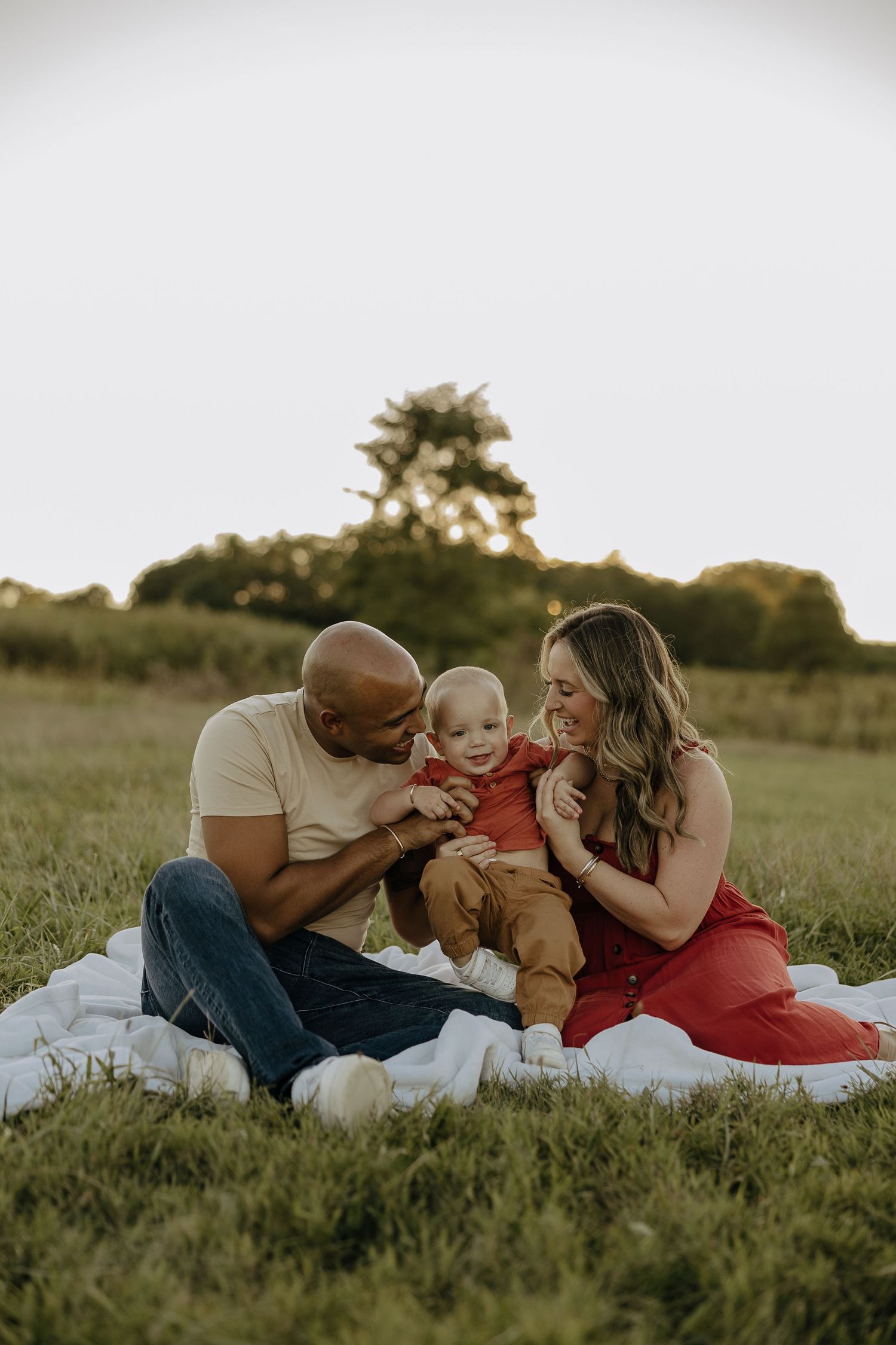 Family shares intimate moments while lying on blanket in grassy field at sunset.