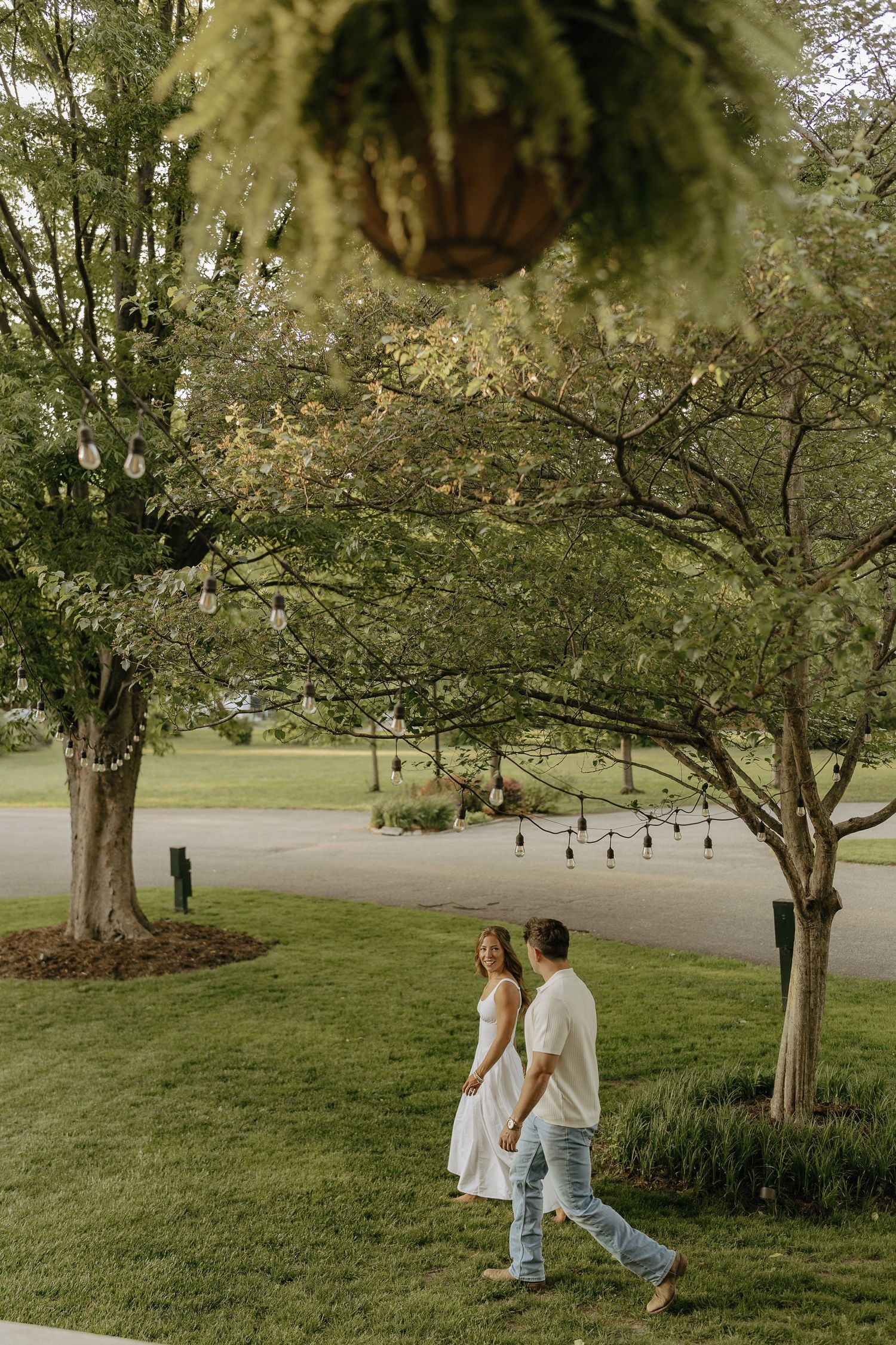 Couple walking hand in hand through lush green yard with string lights and trees at dusk.