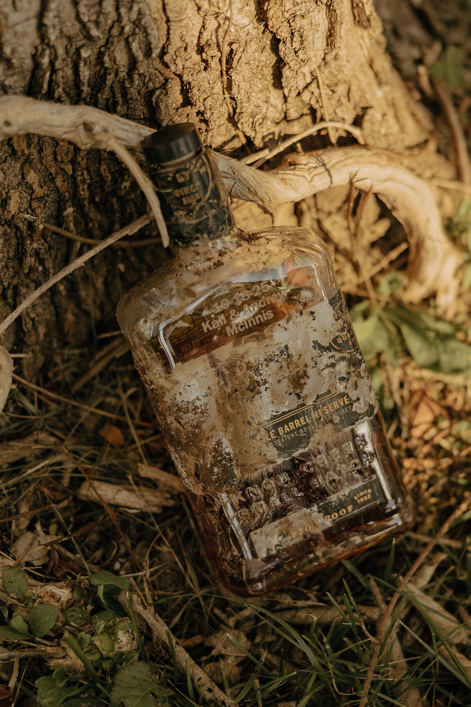 Weathered old glass bottle covered in dirt and debris lying on forest ground among leaves and tree roots.