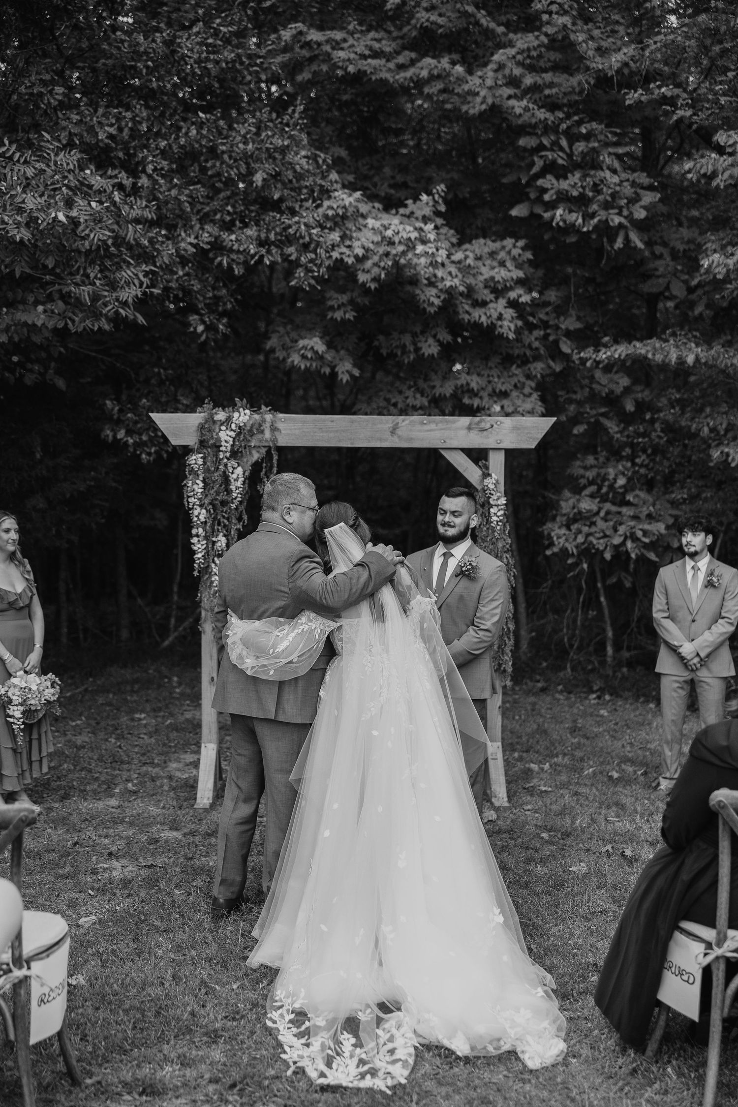 Black and white photo of wedding ceremony under wooden arbor with guests watching.