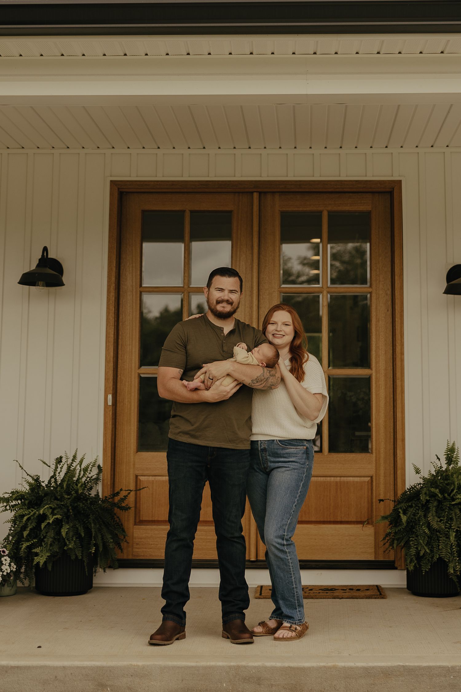 A couple poses casually on their modern farmhouse front porch.