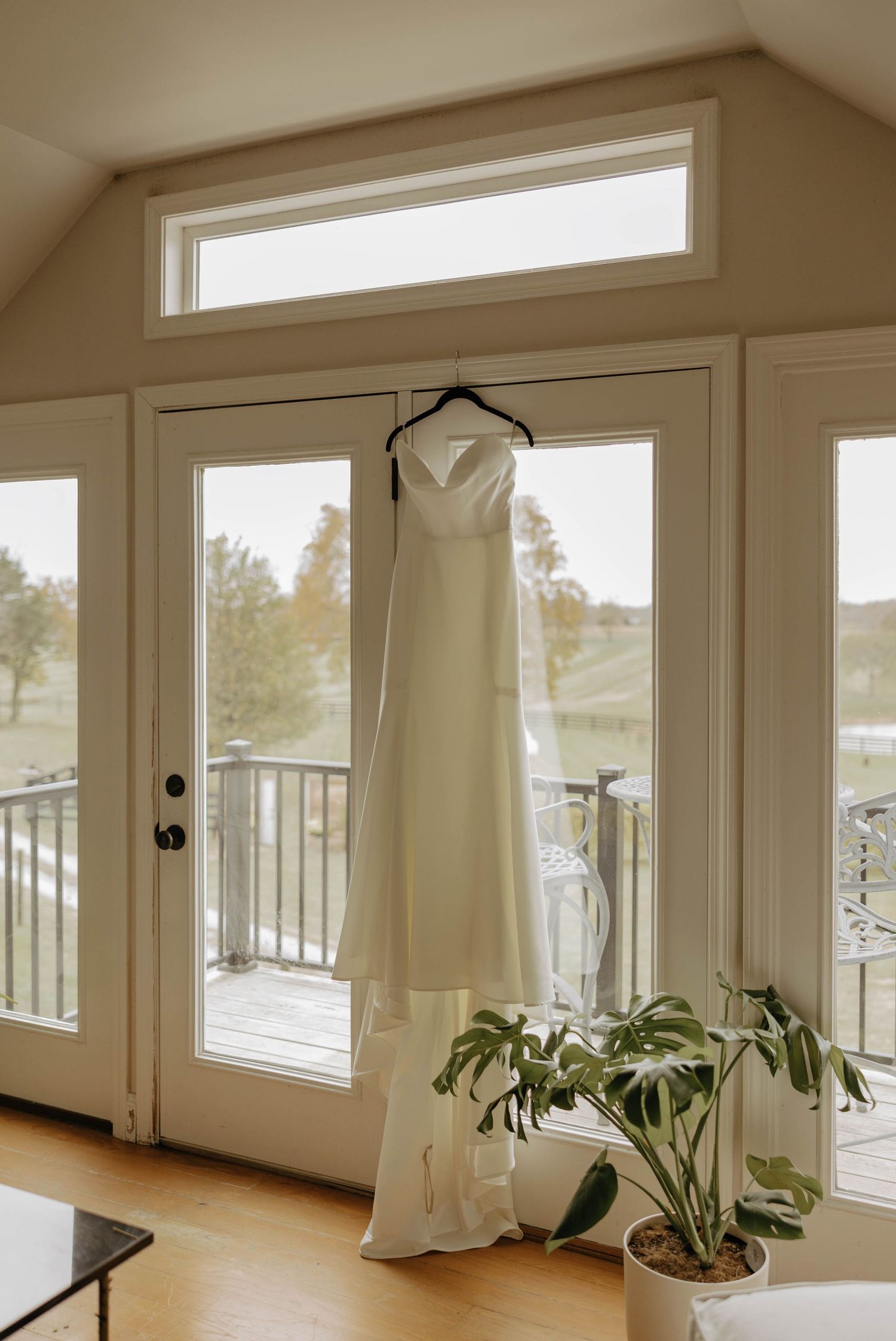 A white wedding dress hangs elegantly in front of large windows with a potted plant below and scenic outdoor view.