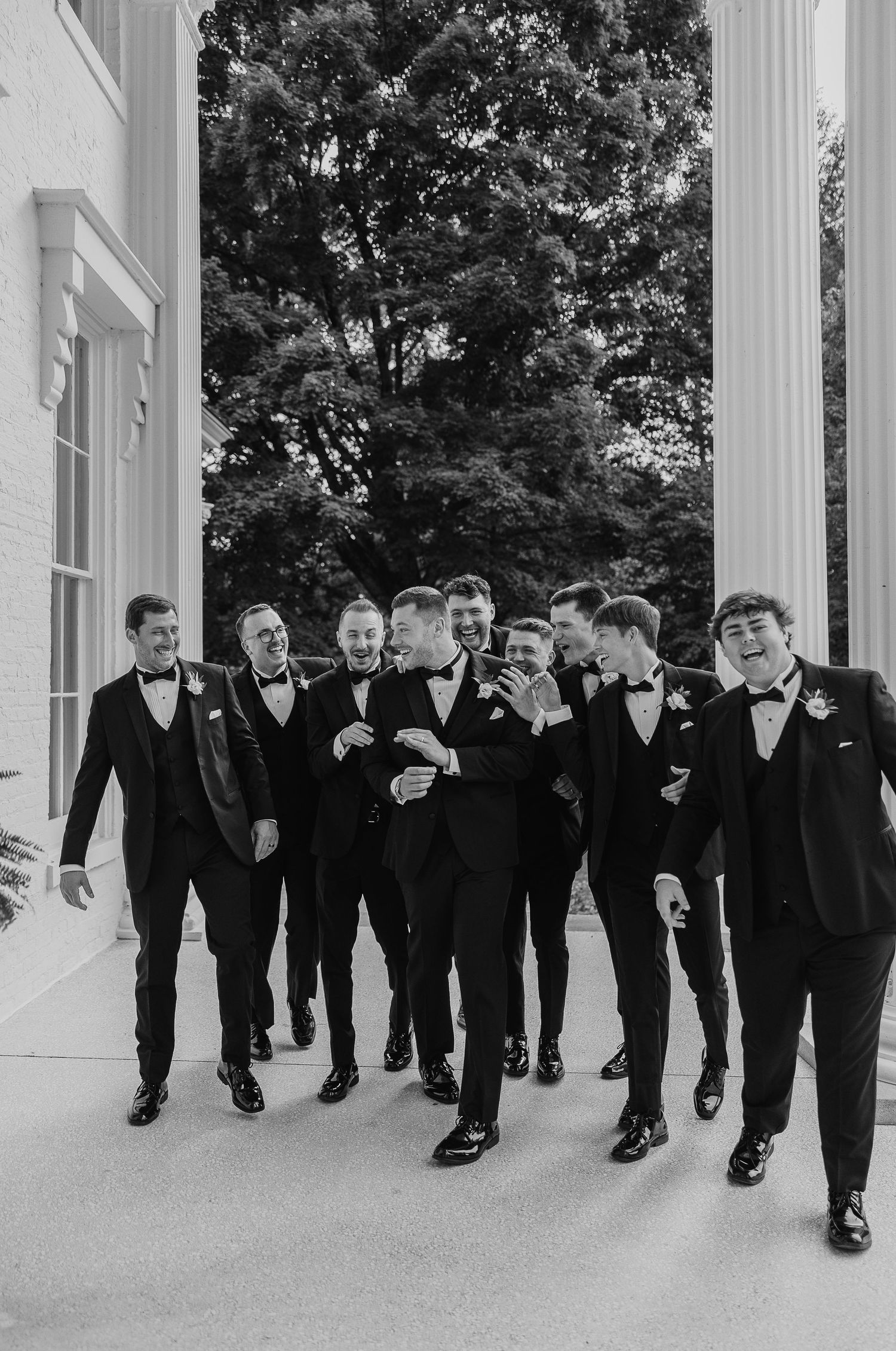 Groom and groomsmen in black tuxedos laughing together between white columns outdoors.