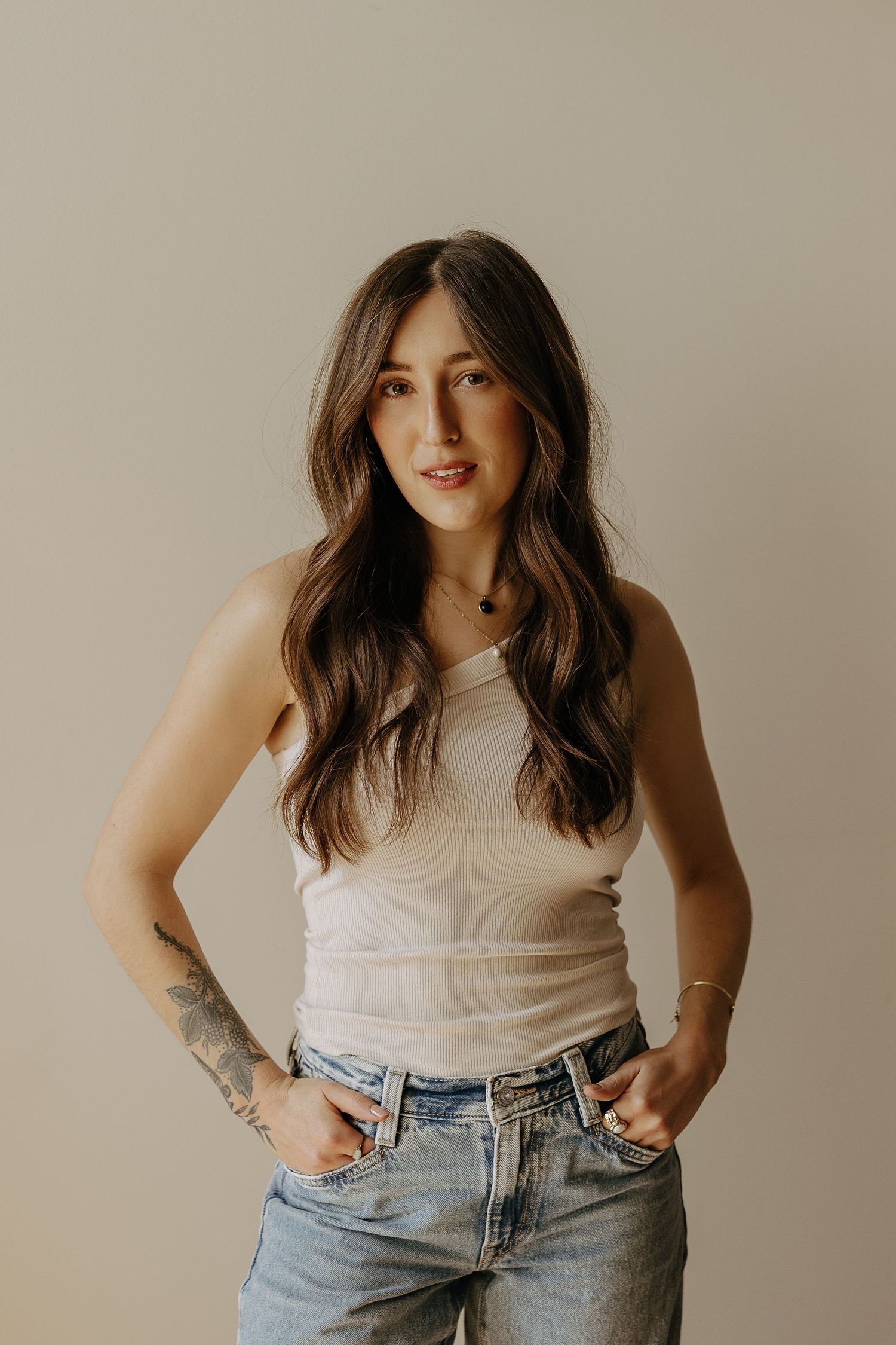 Young woman with long brunette hair in a beige tank top and jeans posing in a studio.