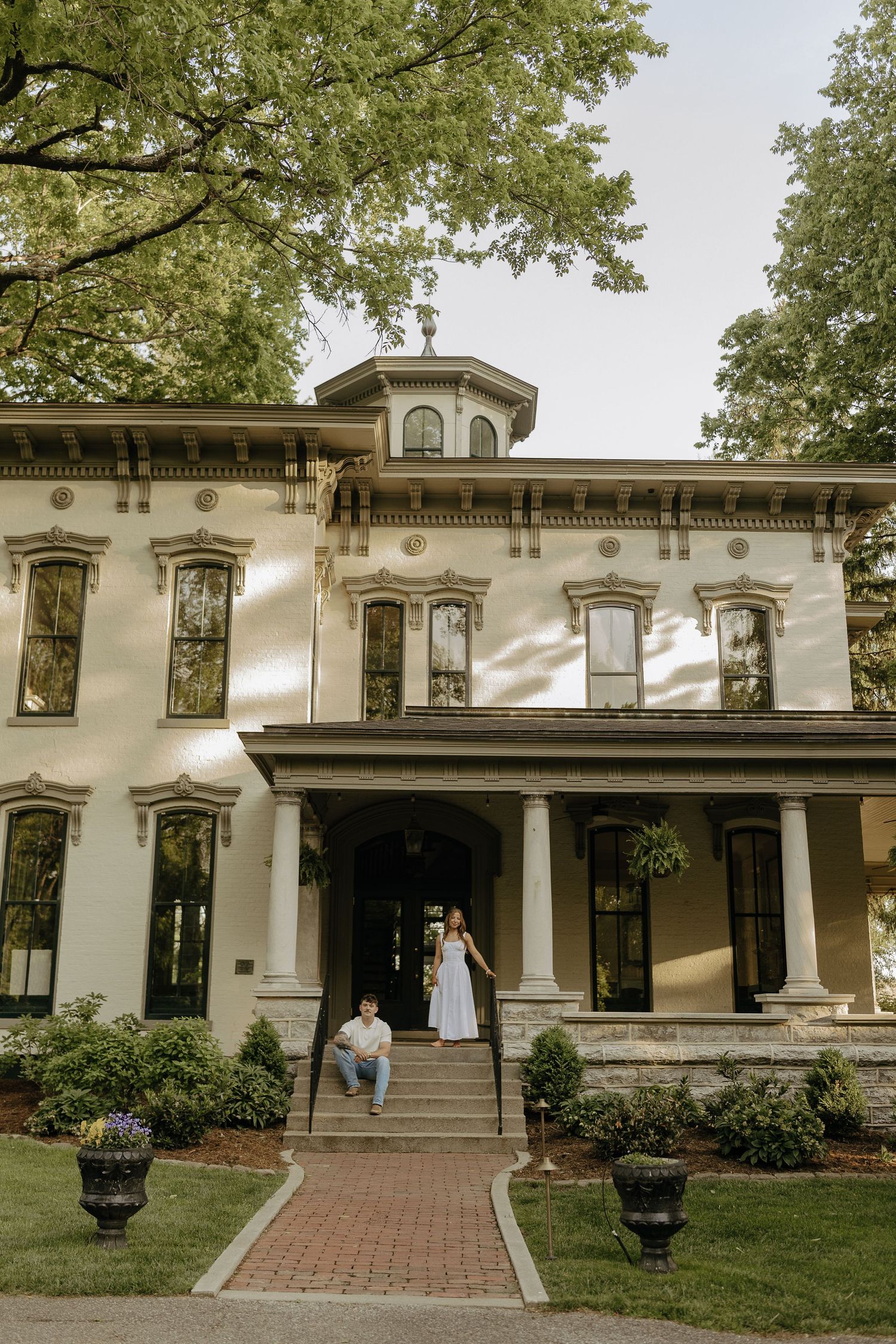 Couple posing on steps of elegant historic white mansion surrounded by lush trees.