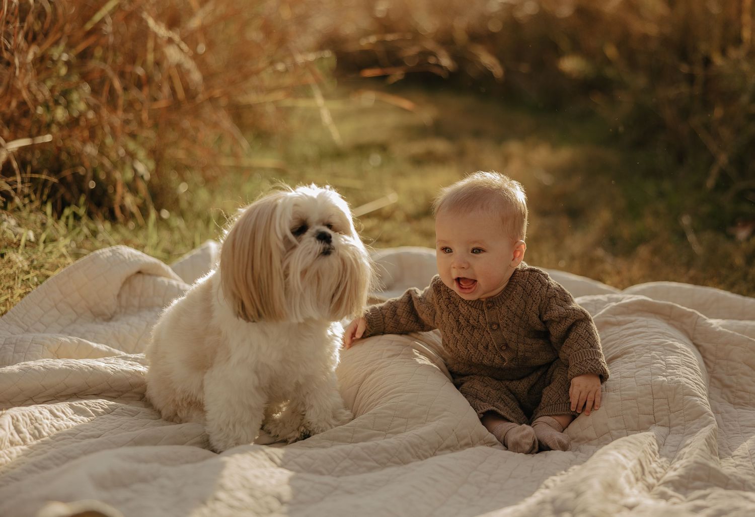 Baby and white dog lying together on white blanket outdoors.