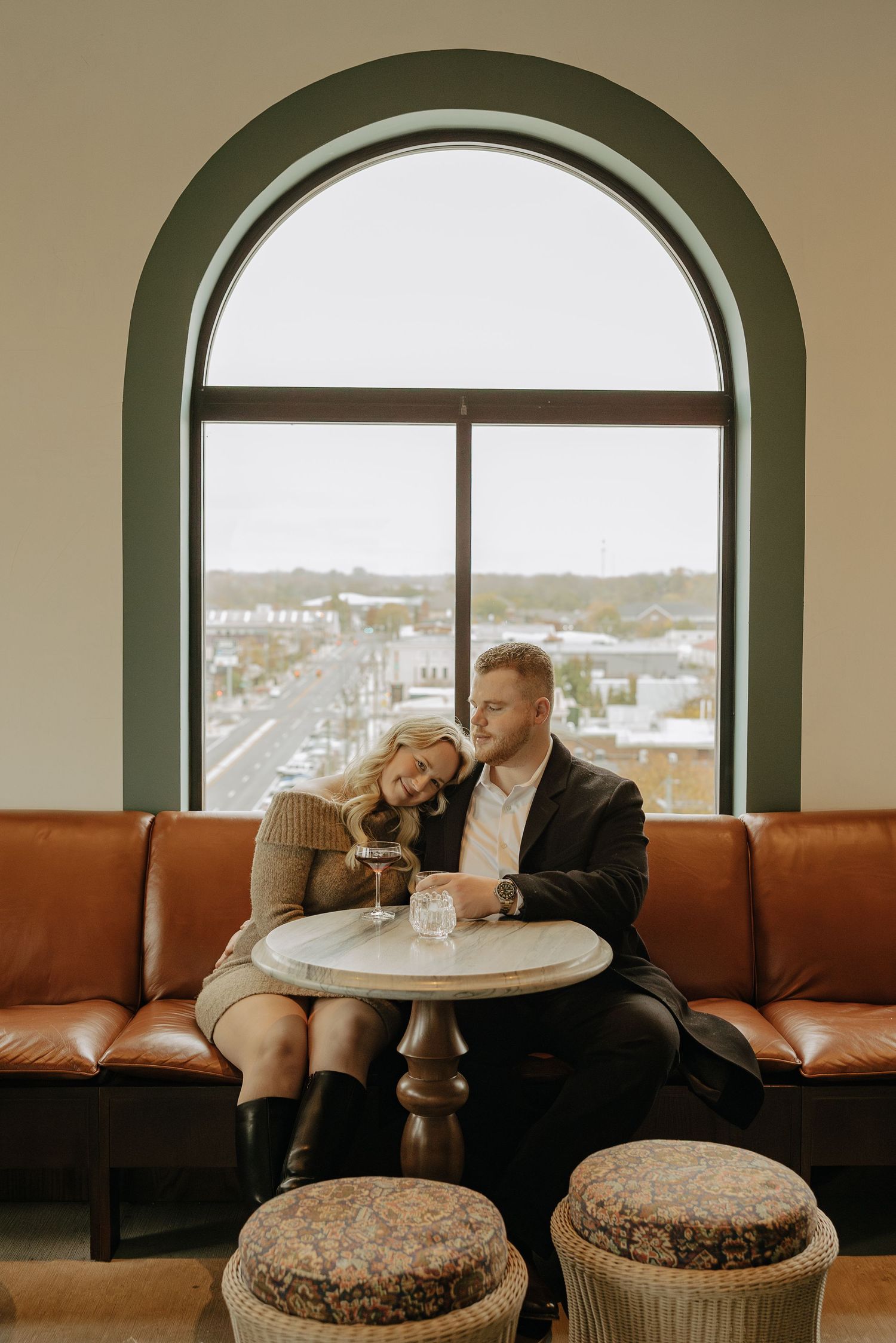 A couple sits together on a brown leather couch beneath arched windows overlooking a city view.