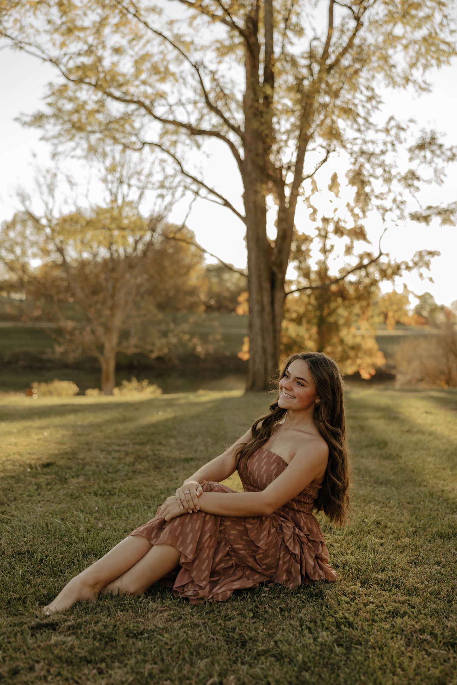 A person in a brown dress sits gracefully on grass during golden hour.