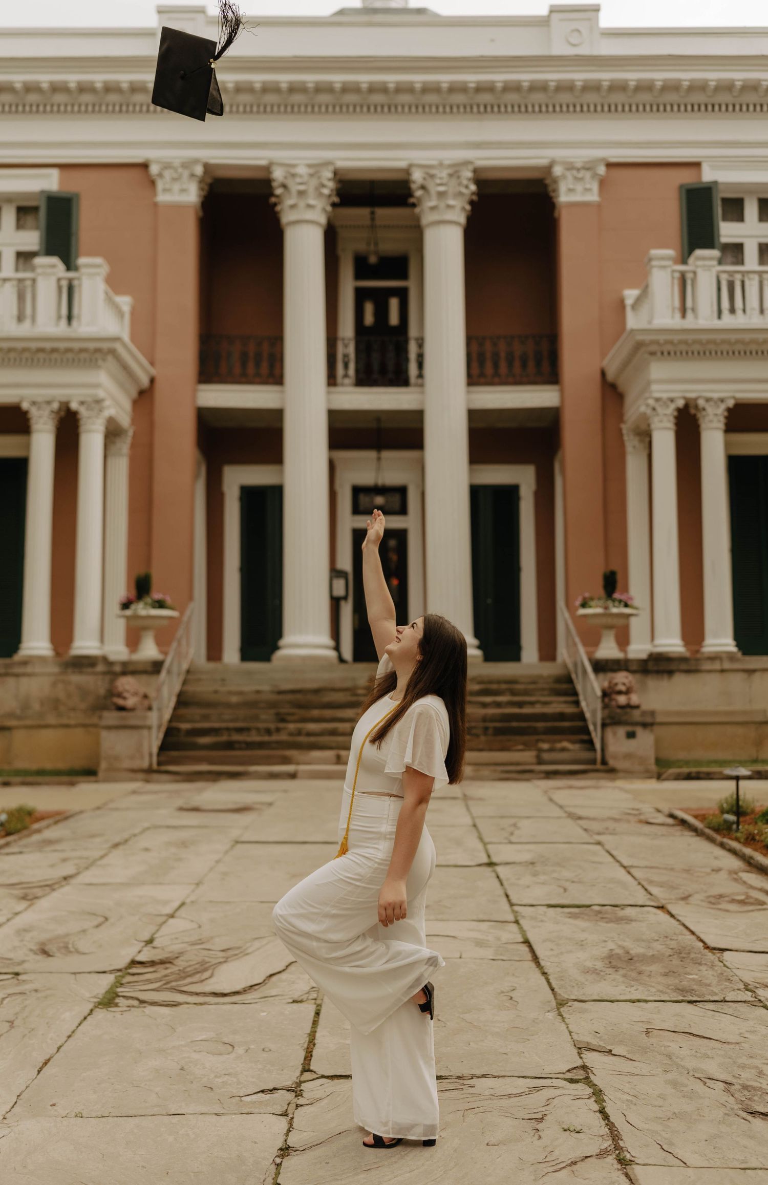A graduate twirls in a white dress against the backdrop of a classical building with columns.