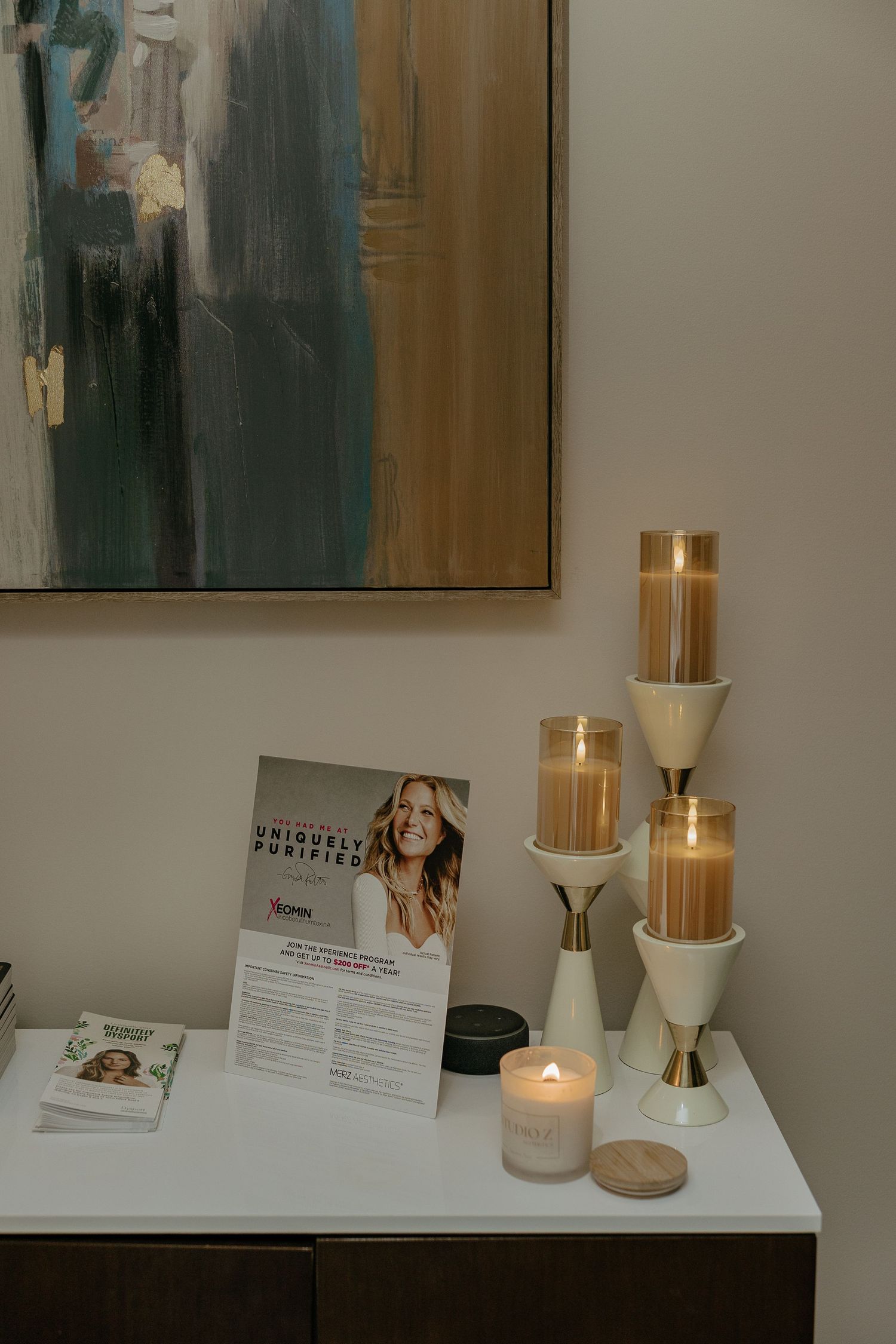 Elegant display of beauty products and candles on white counter with artwork in background.