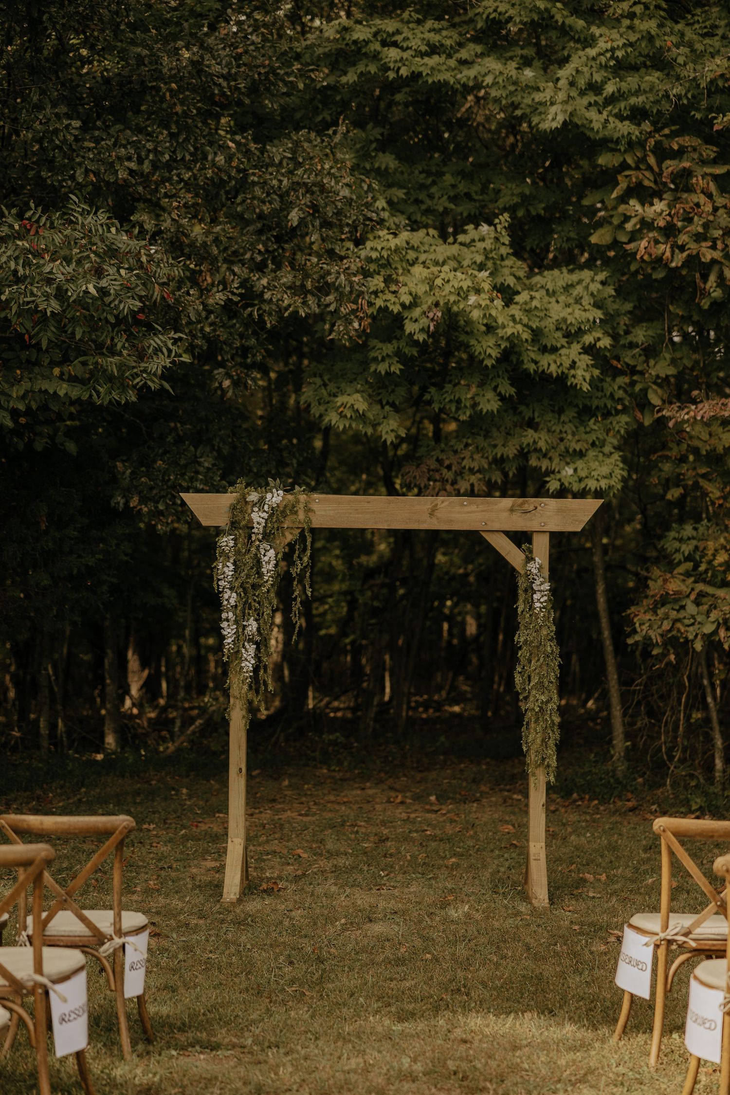 A rustic outdoor wedding ceremony setup with wooden arbor and white chairs in a wooded area.