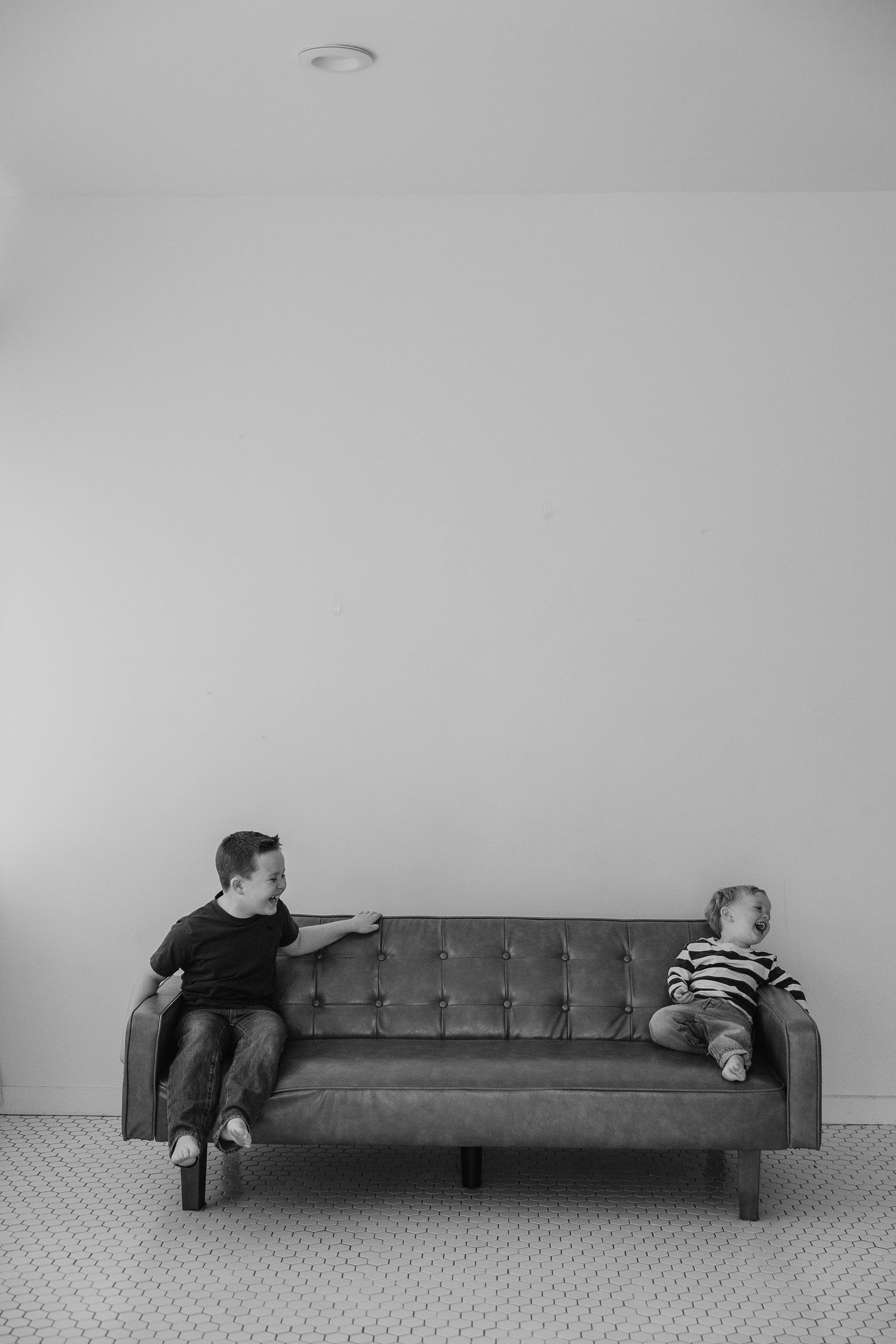 Two children sit at opposite ends of a gray tufted couch in minimalist black and white photo.