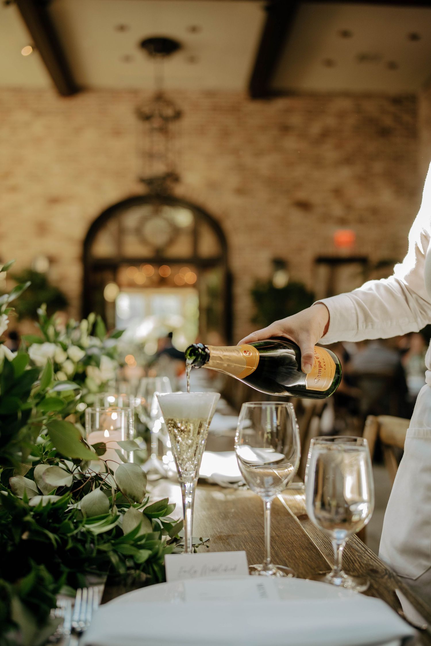 A server pours wine at an elegant table setting decorated with greenery and glassware at a rustic venue.