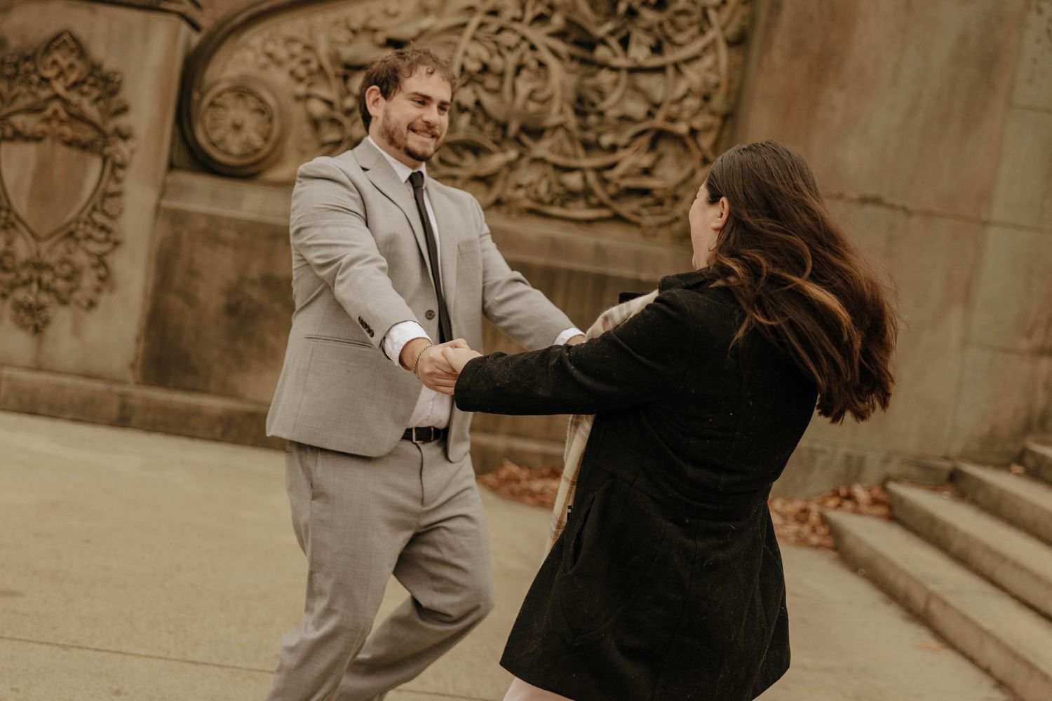 A couple dances playfully together on stone steps in front of an ornate architectural backdrop.