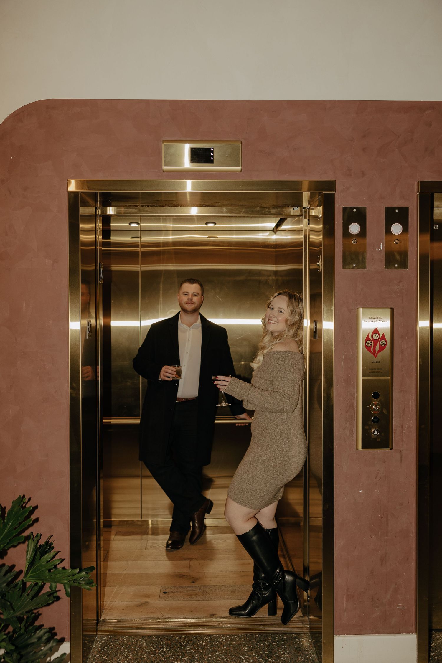 Stylish couple posing playfully in a modern elevator lobby with gold accents.